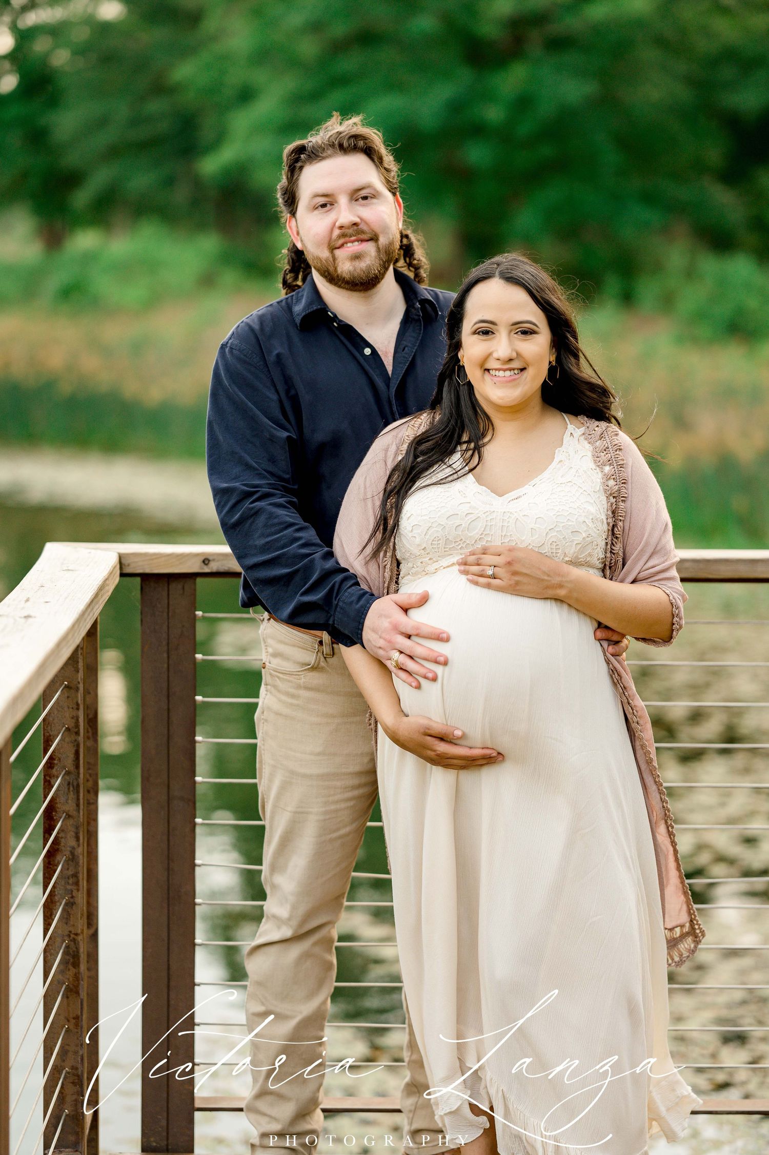 couple taking maternity photos wearing blue button down shirt and wife wearing white lace dress for a sunset photo