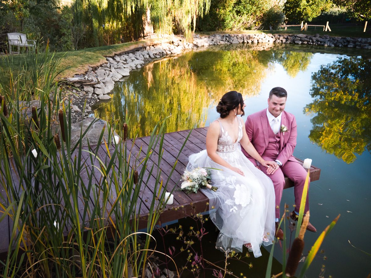 A couple in wedding attire sits on a wooden dock overlooking a tranquil pond surrounded by lush greenery at sunset.