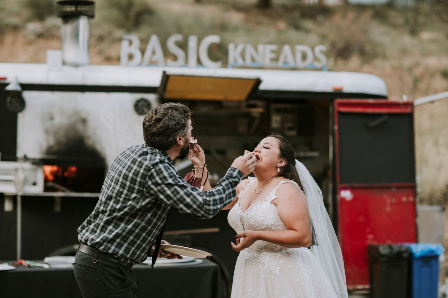 Bride and groom feeding each other slice of pizza while wearing wedding attire