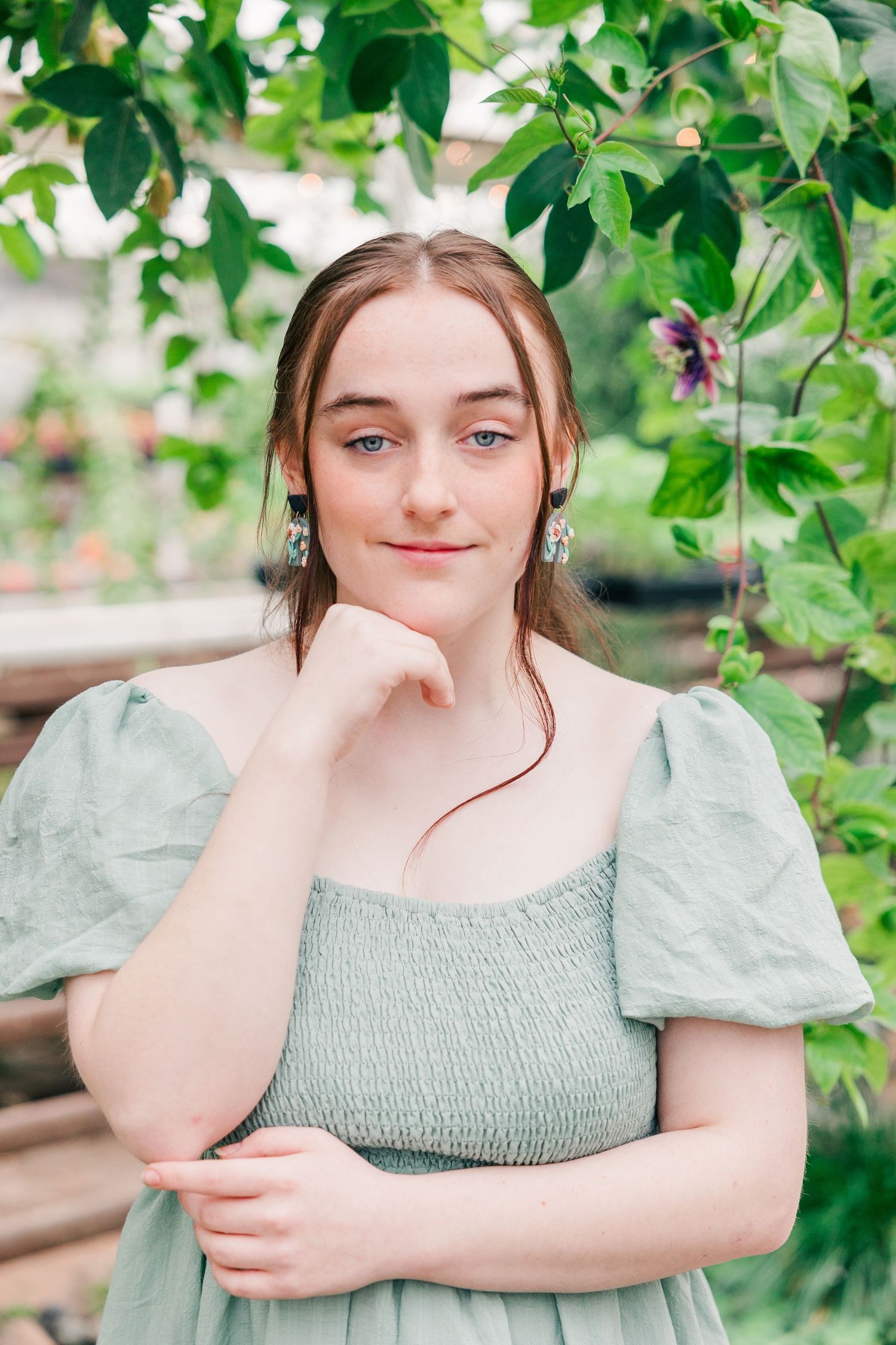 Portrait among green foliage wearing sage dress with puffed sleeves and delicate earrings.