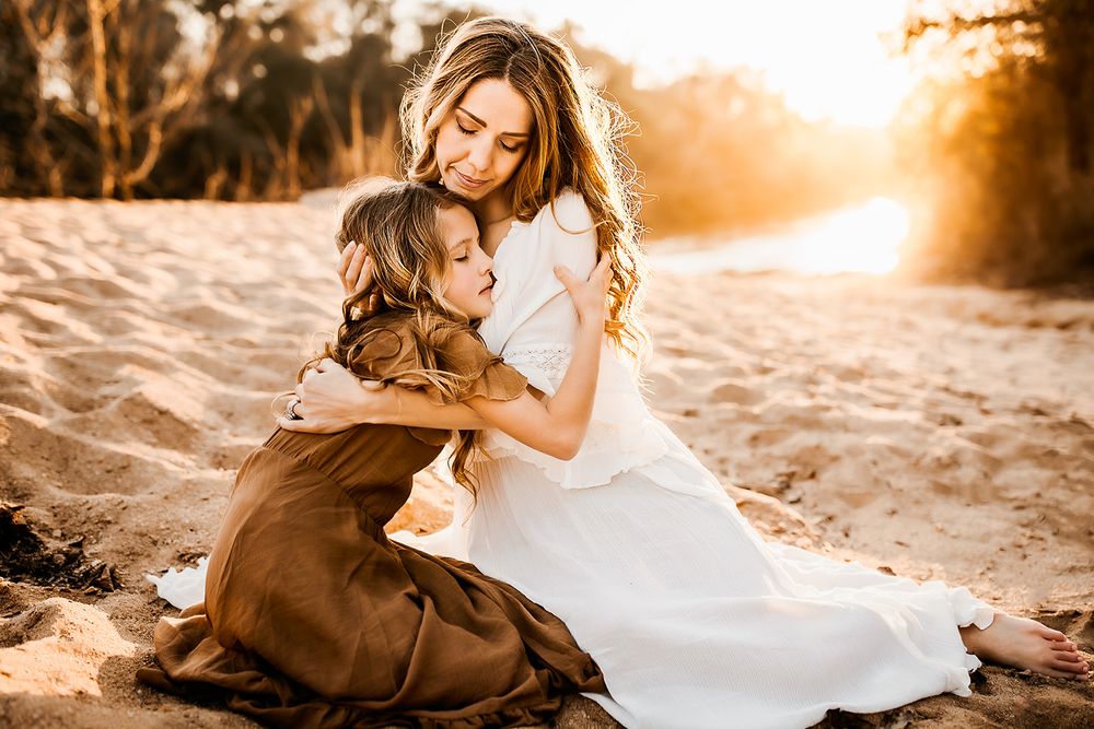 A mother and daughter hug while sitting on a sandy beach during golden hour.