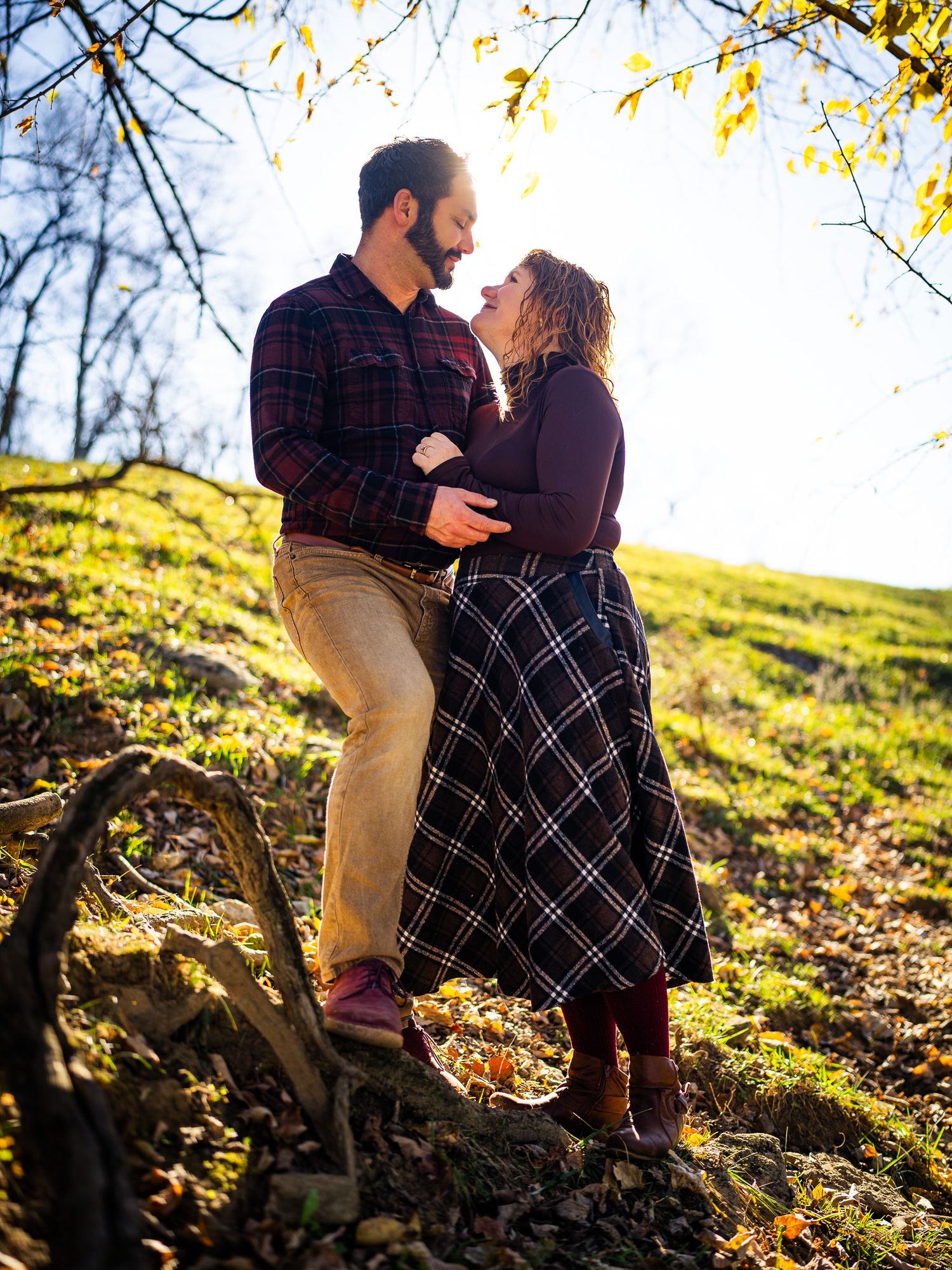 A couple embraces on a hillside during autumn with yellow leaves overhead.