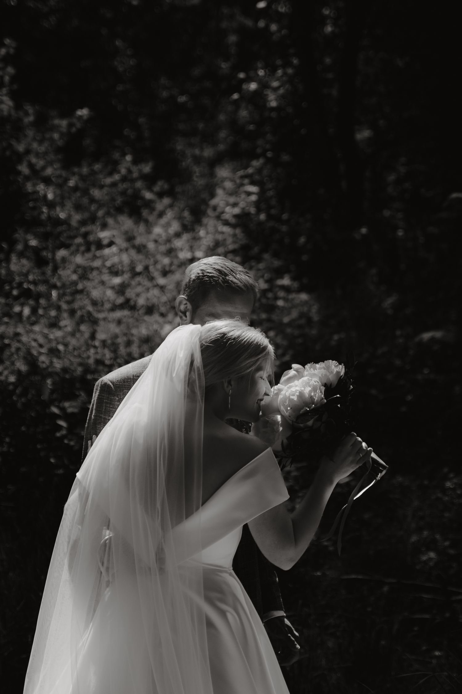 Intimate black and white wedding portrait with flowing veil against dark background.