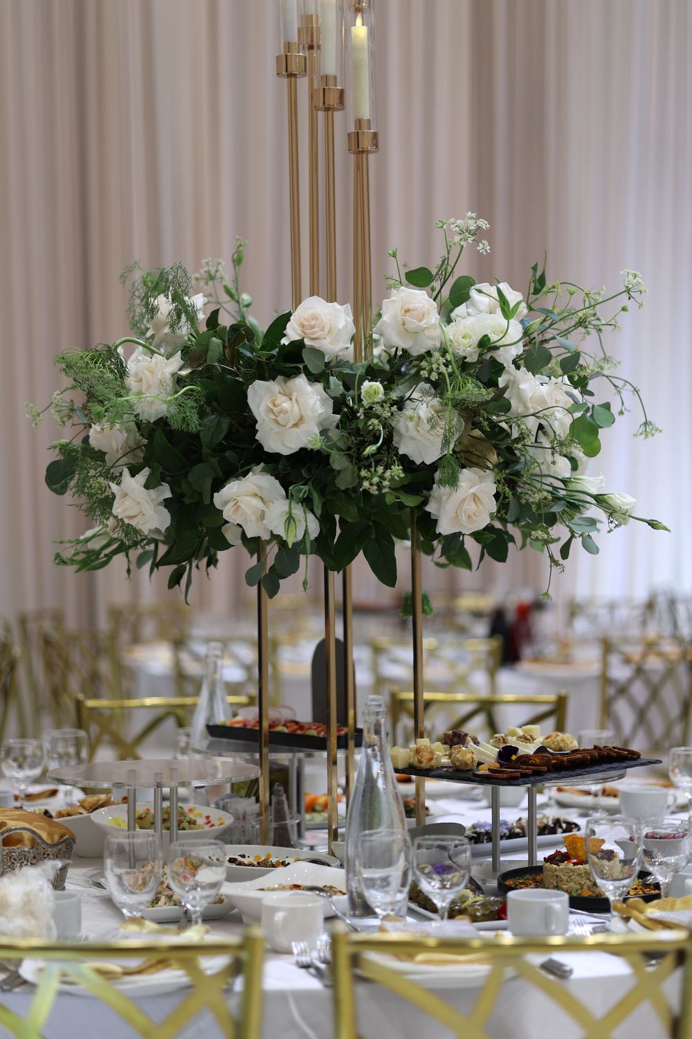 Elegant white floral centerpiece with roses and greenery on tall gold stand adorning formal dining table setting.