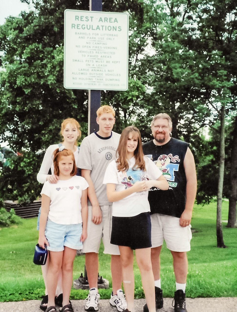 A family group photo taken at a rest area with regulations sign visible in the background.
