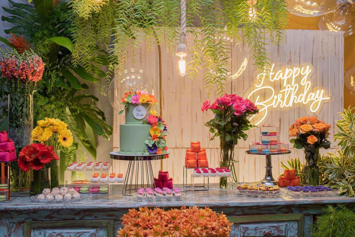Elegant birthday party dessert table setup with floral decorations, cake stands, and glowing 'Happy Birthday' sign on rustic backdrop.