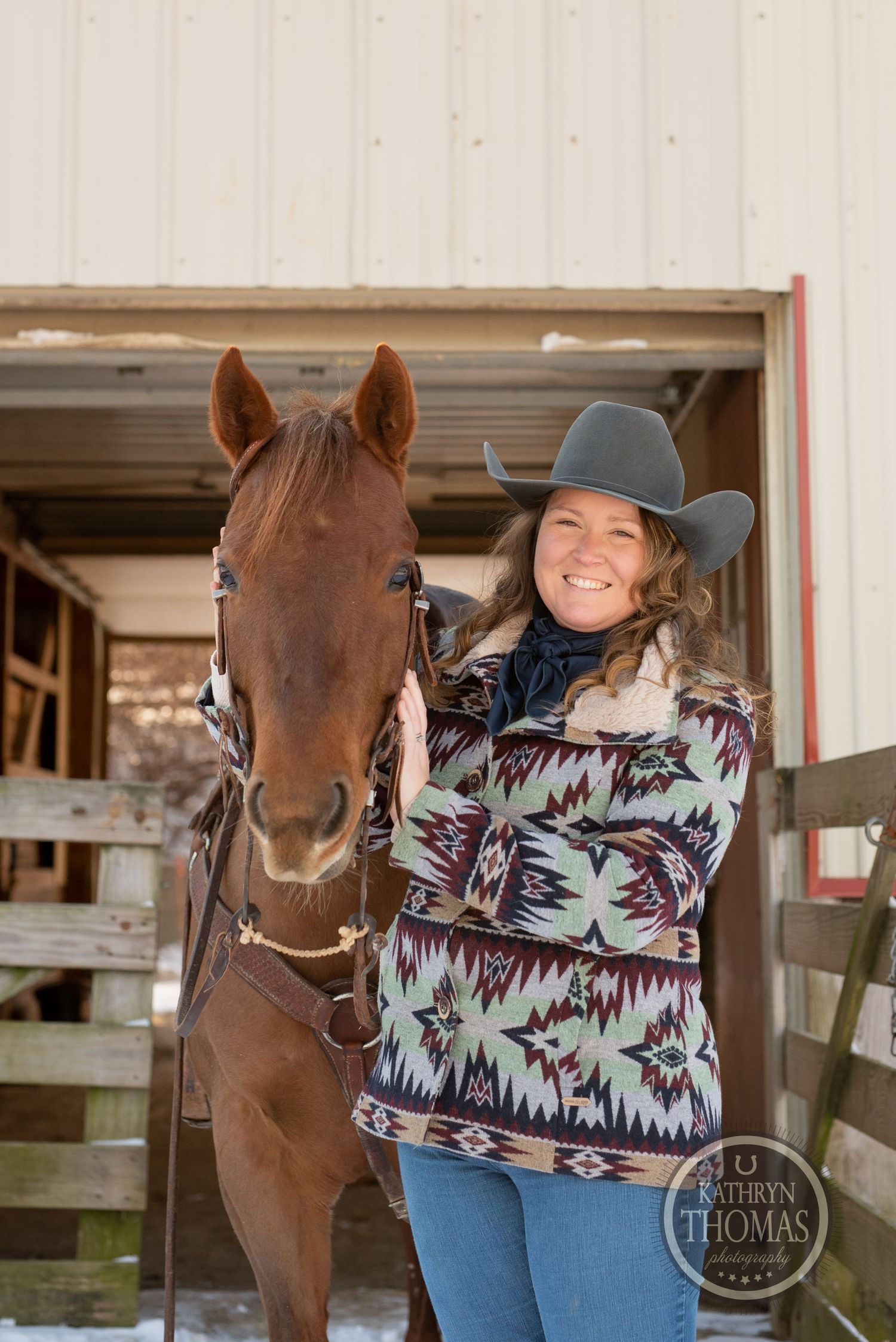 Handler in patterned sweater poses with chestnut horse at stable entrance.