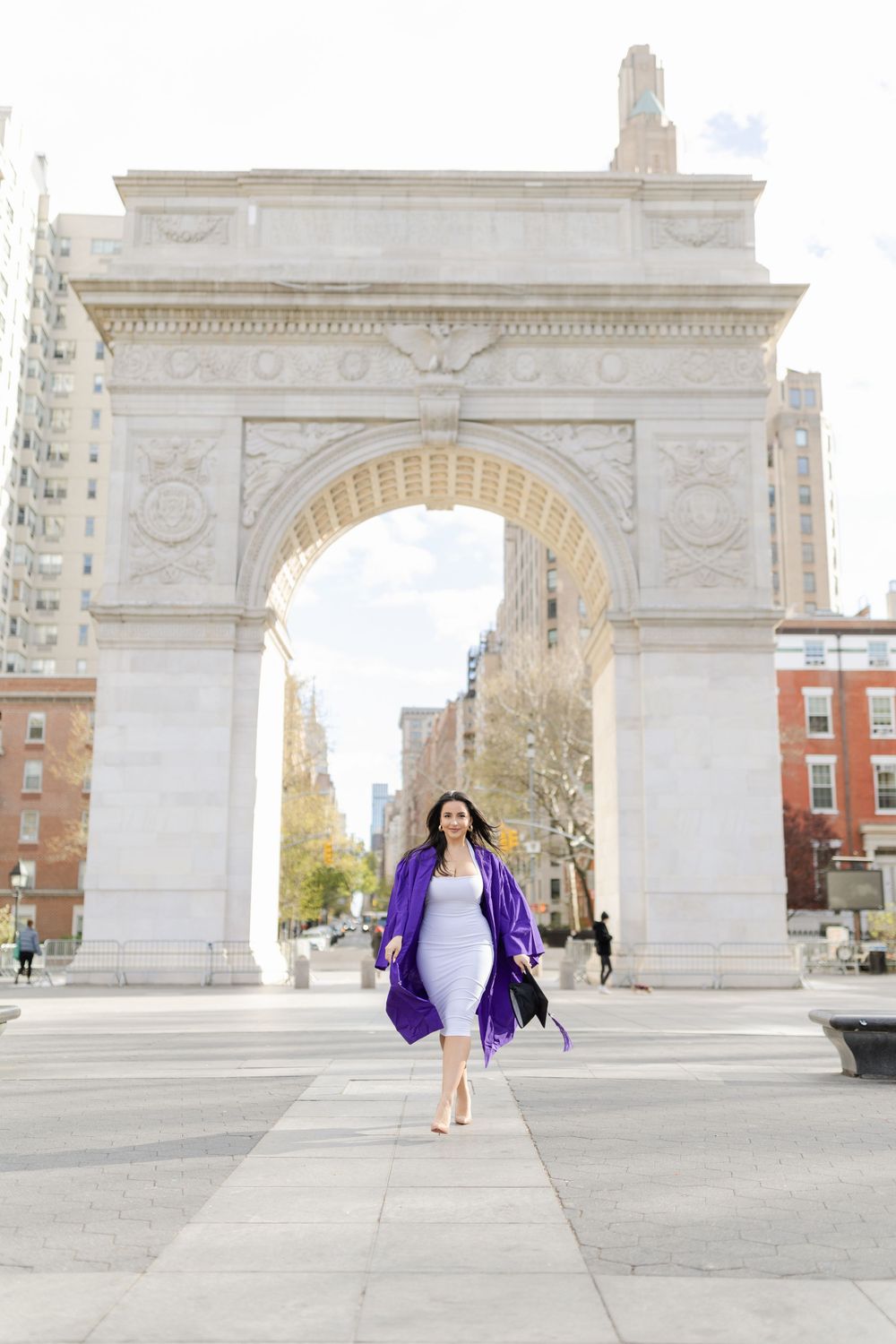 Graduate in white dress and purple robe walks confidently through Washington Square Arch in New York City.