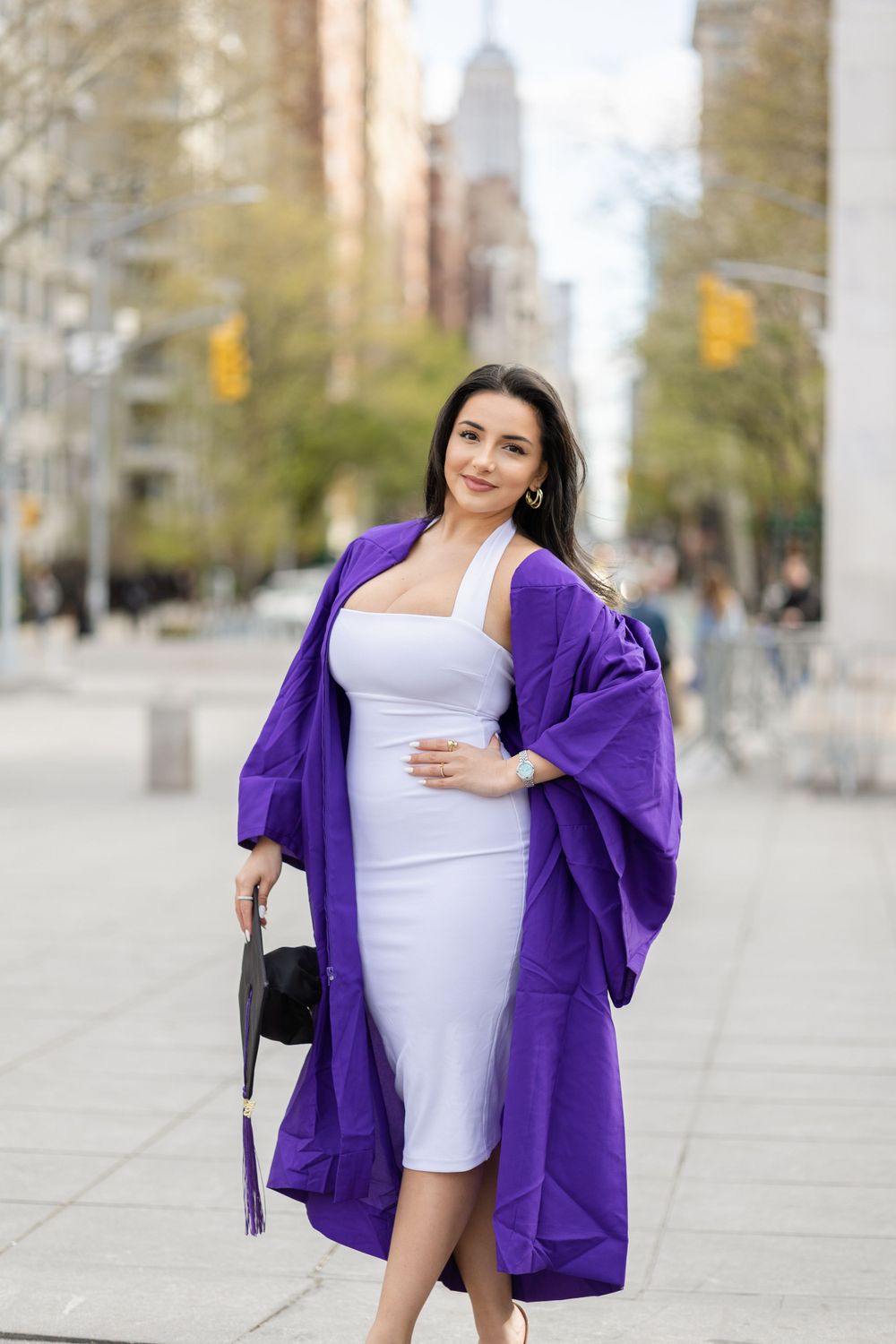 Graduate in white dress and purple graduation gown poses on city sidewalk with skyscraper backdrop.