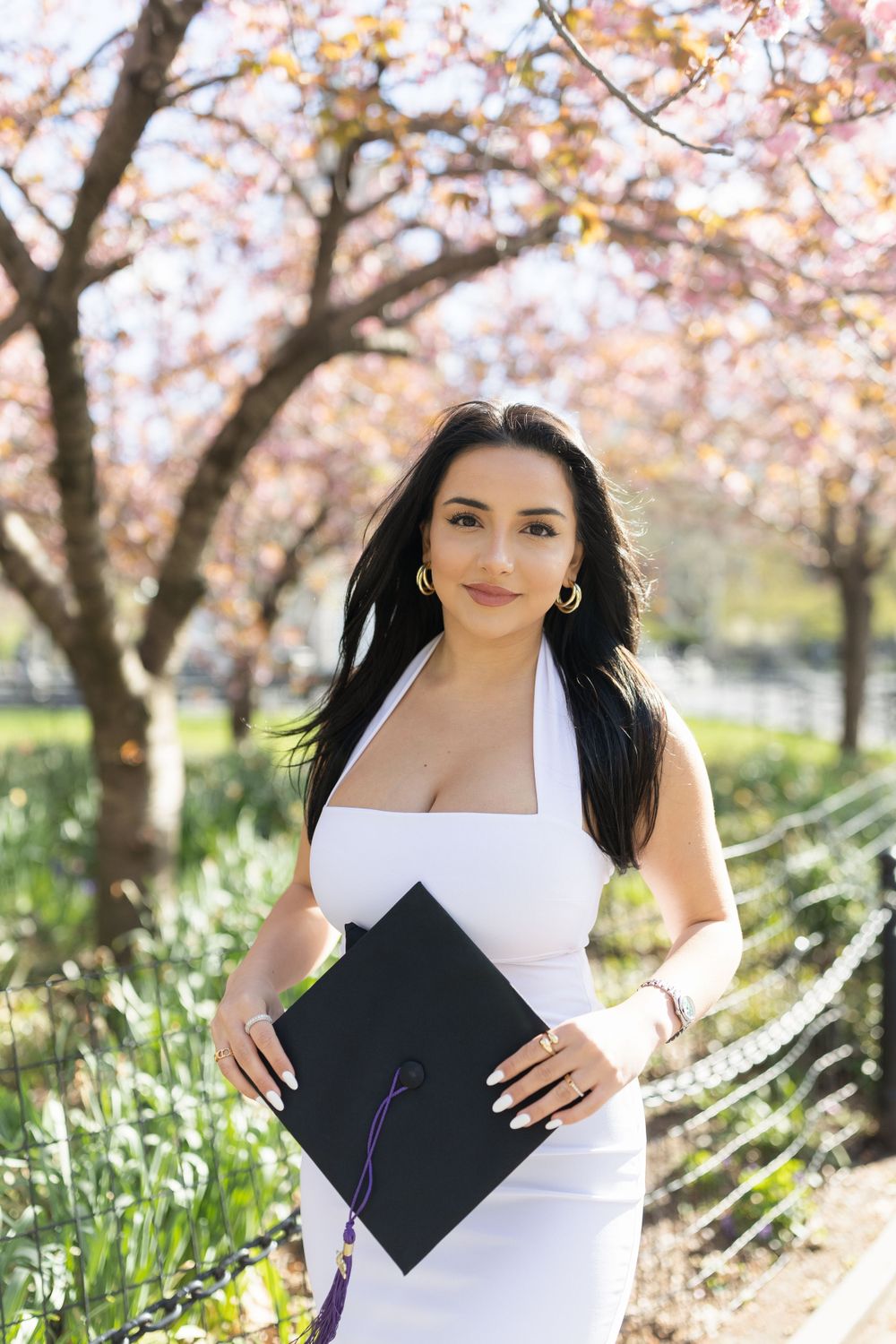 Graduate in white dress holds diploma while posing under blooming cherry blossom trees.