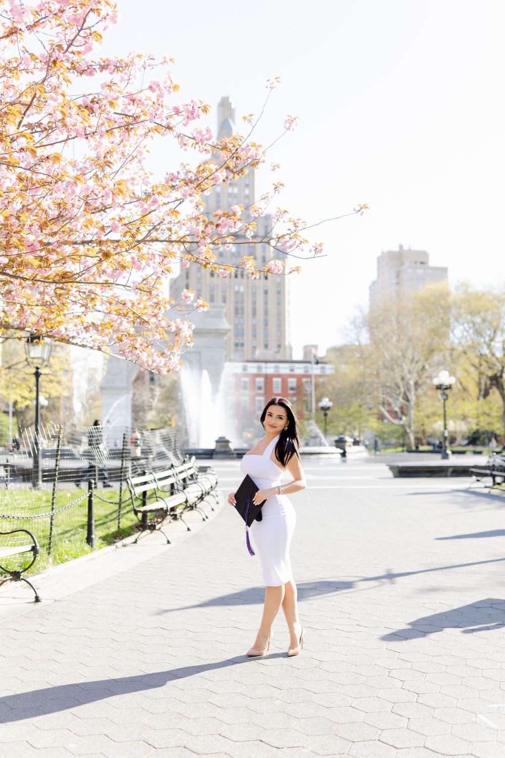 Graduate in white dress poses with cherry blossoms and Washington Square Park fountain in background.