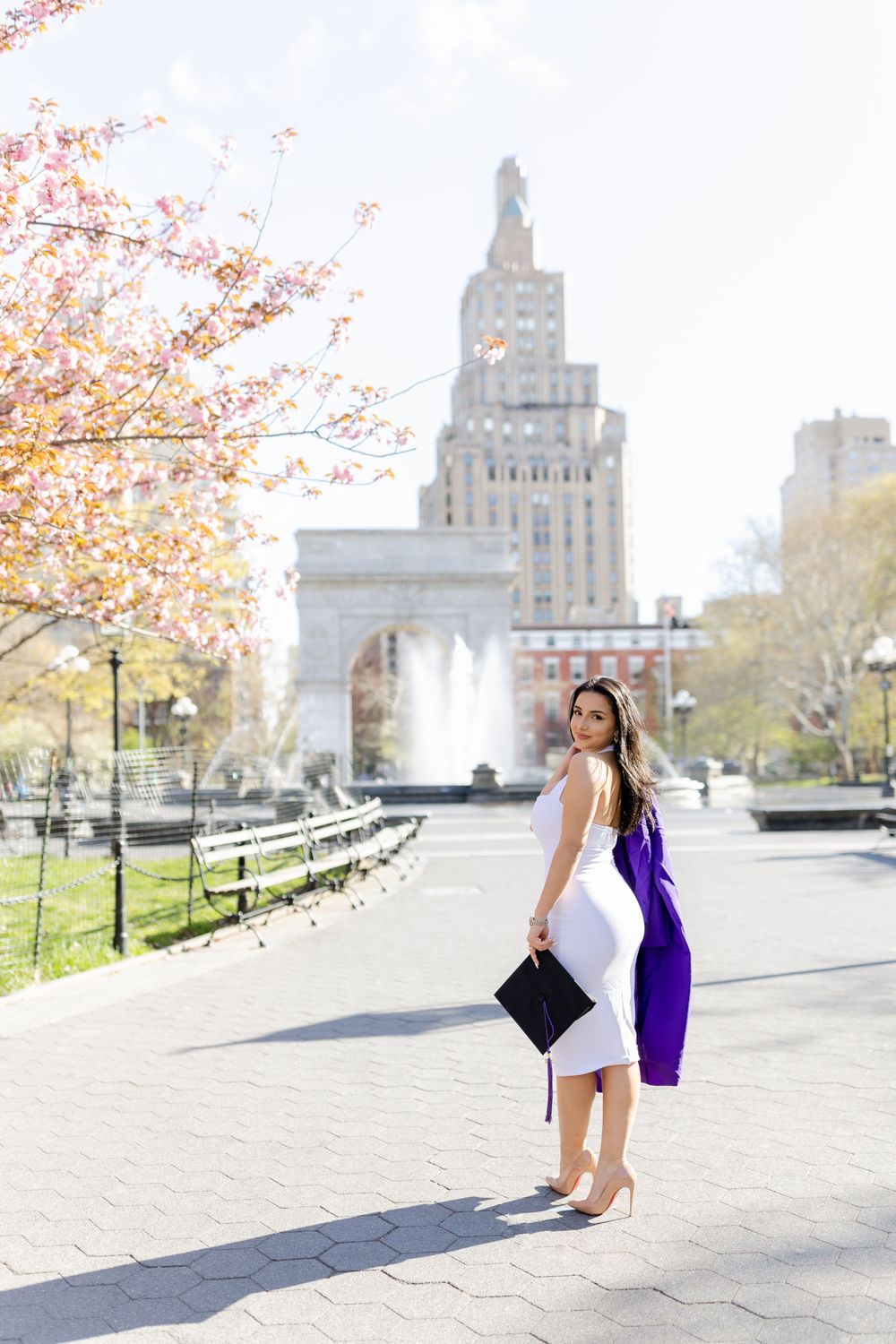 Graduate poses with diploma and purple gown against iconic Washington Square Park backdrop.