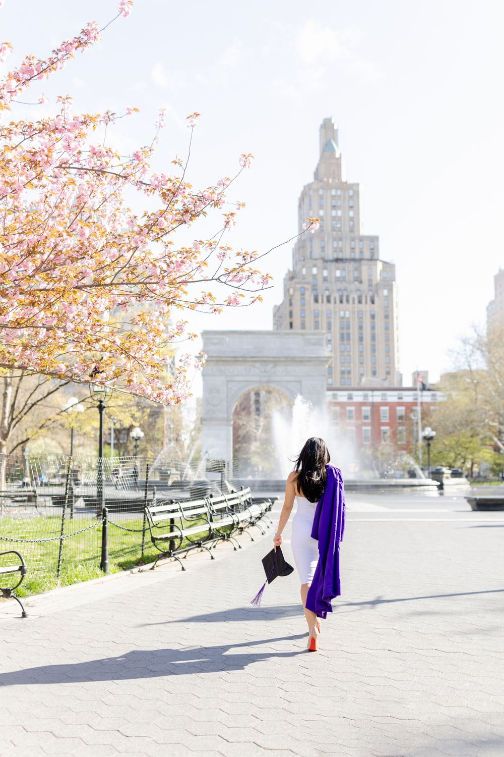 Graduate in flowing purple gown twirls near Washington Square Park on spring day.