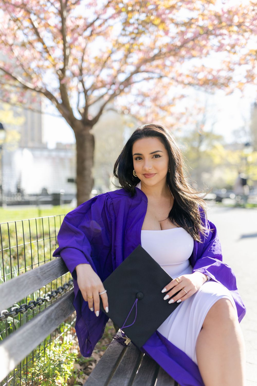 Graduate in purple cap and gown sits gracefully on a park bench under blooming cherry blossoms in spring.
