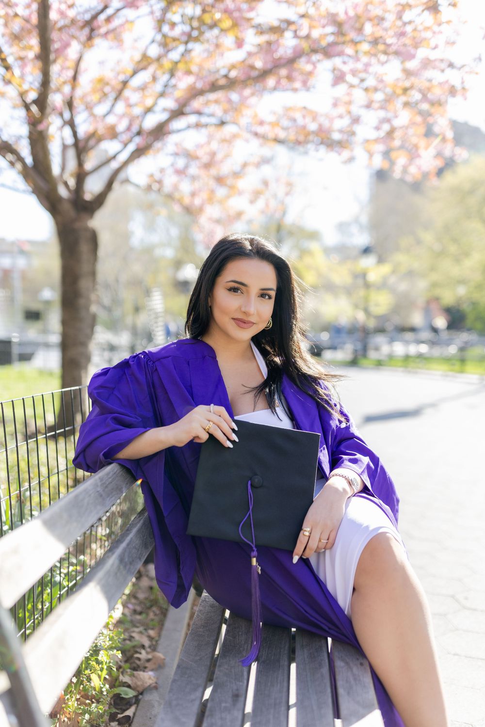 Graduation portrait featuring purple academic regalia and diploma on a wooden bench with spring foliage.