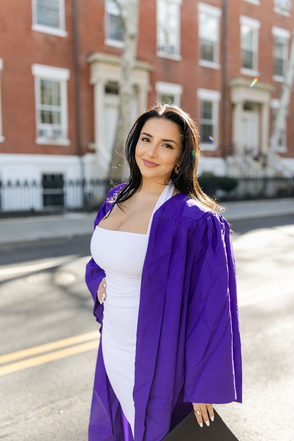 Graduate poses in vibrant purple graduation gown against classic brick building backdrop.