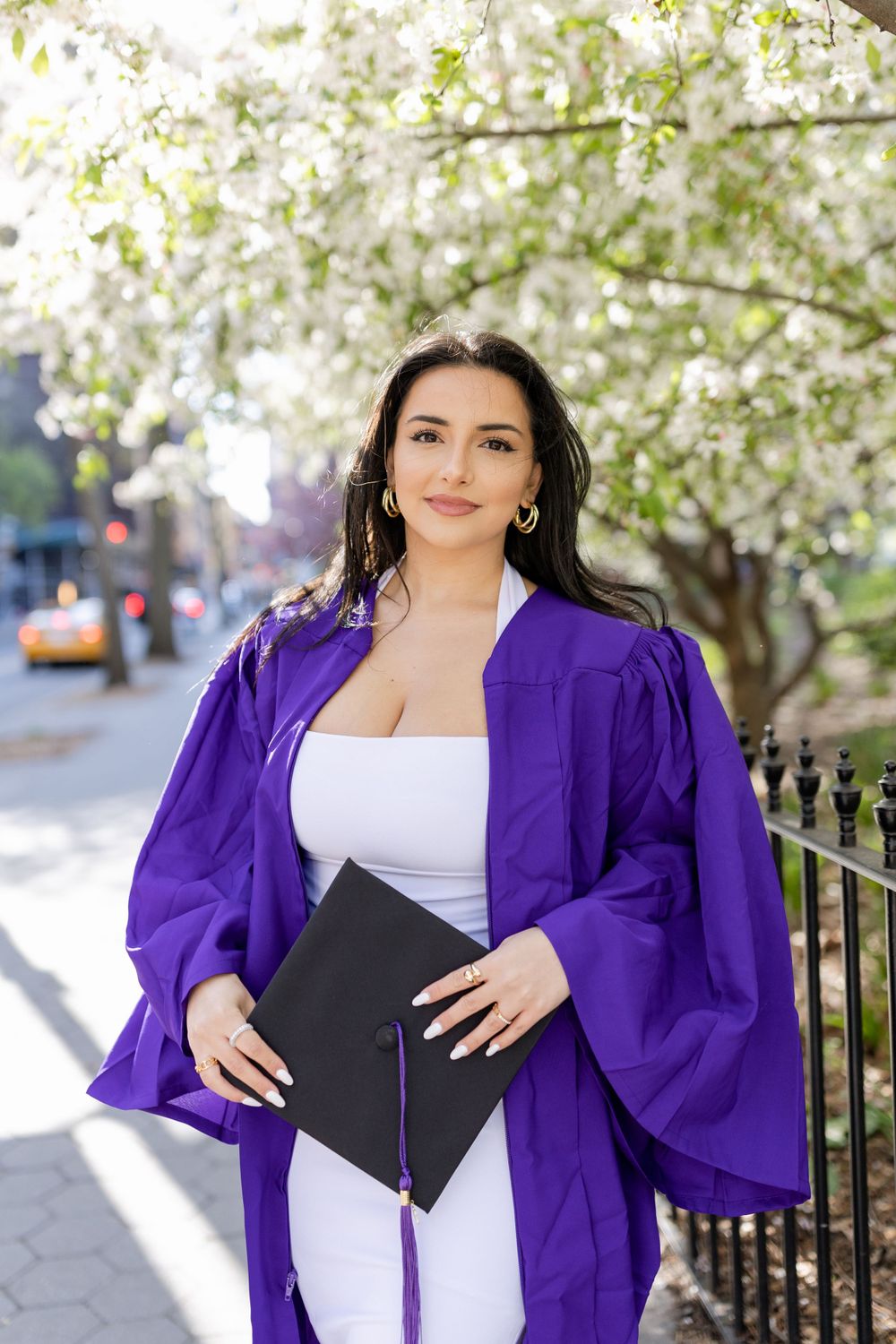 Graduate holding diploma stands beneath white spring blossoms wearing purple graduation regalia.