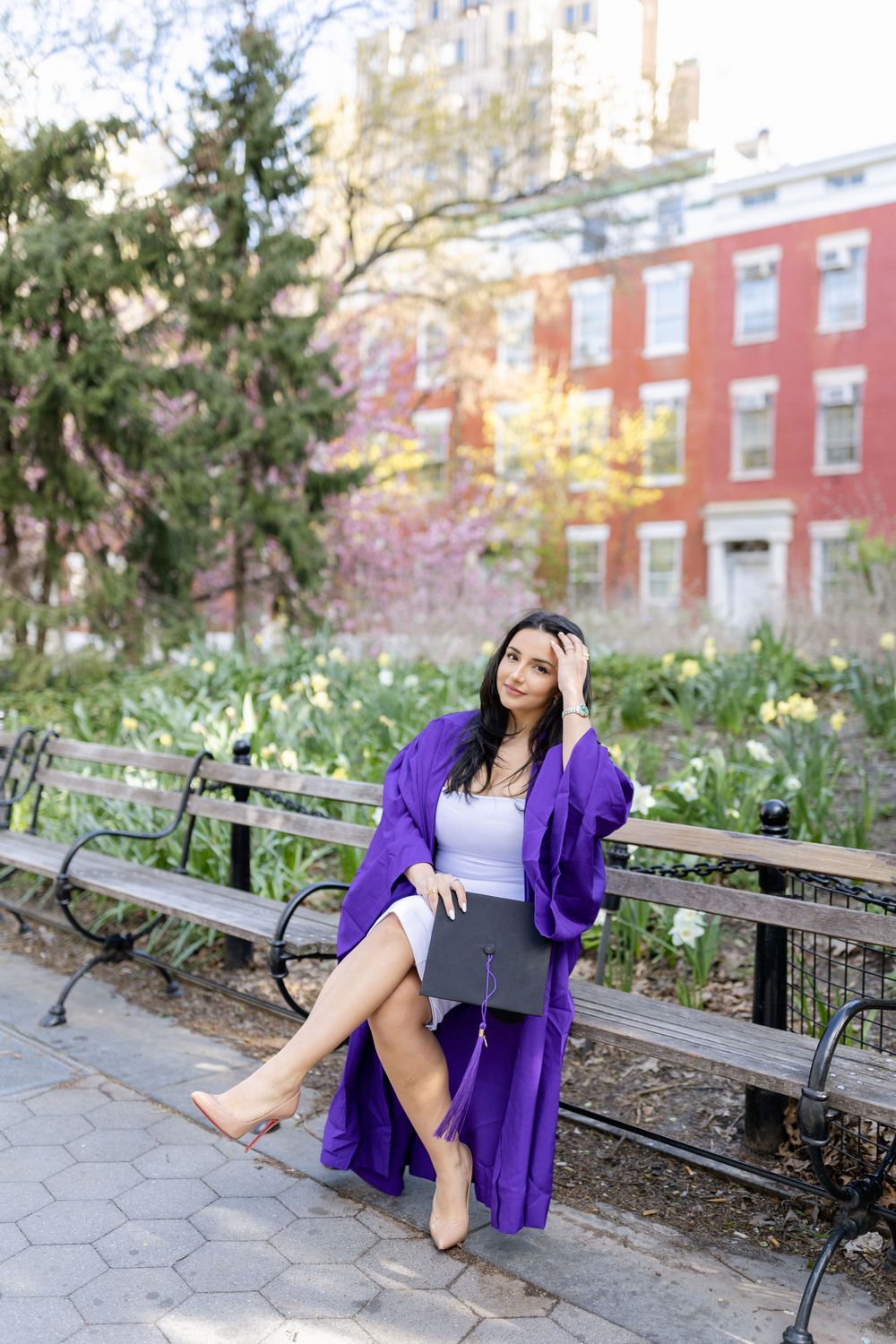 Graduate in purple gown relaxes on park bench holding diploma during spring photo session.