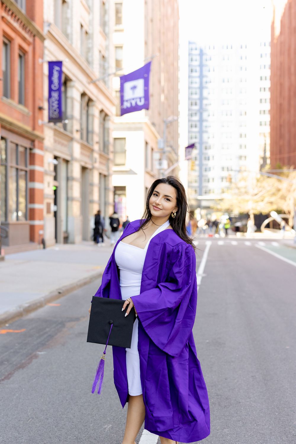 Graduate in purple regalia poses on urban street with historic architecture and NYU banners.