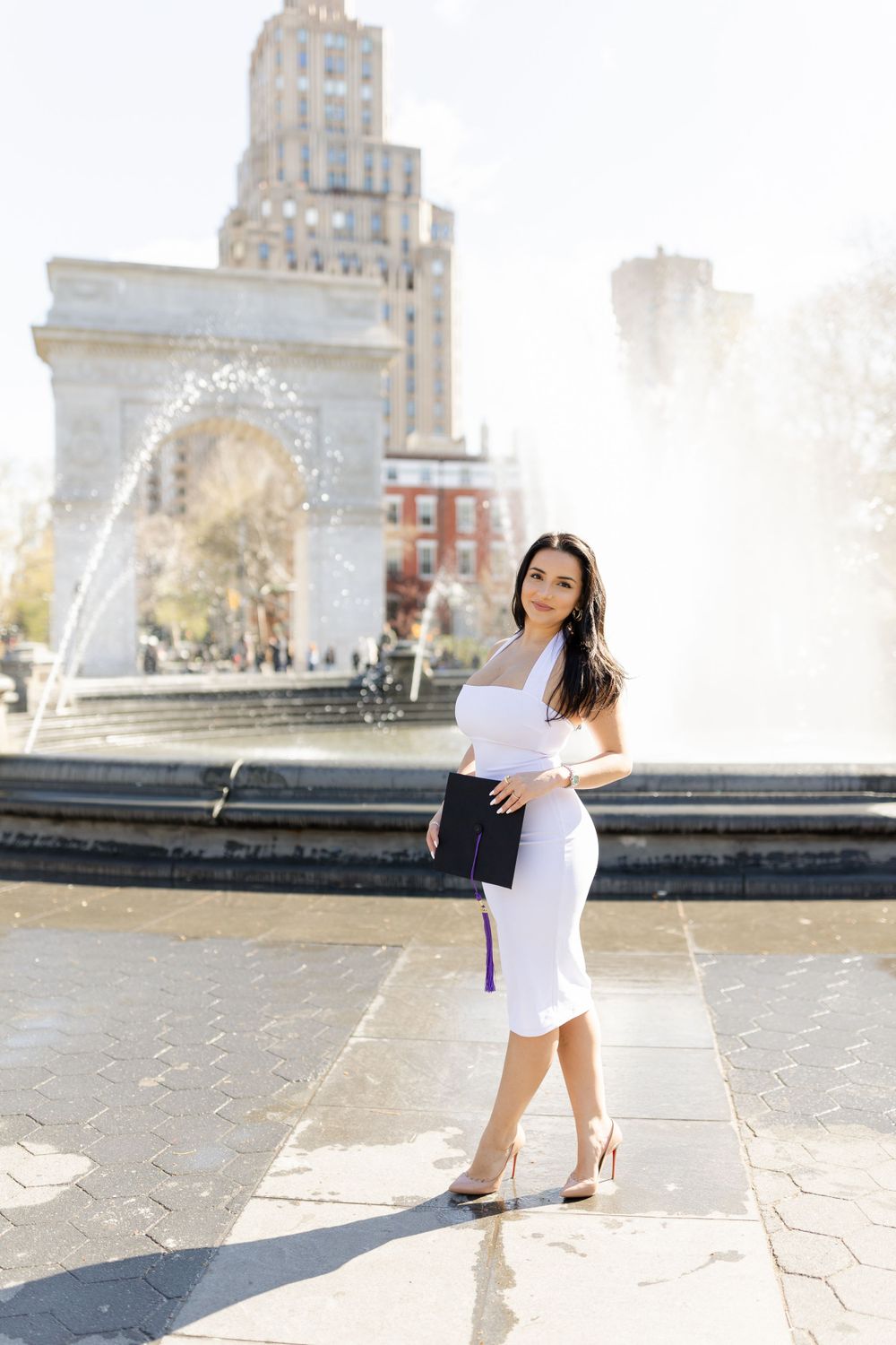 Graduate poses with diploma near Washington Square Park fountain in elegant white dress.