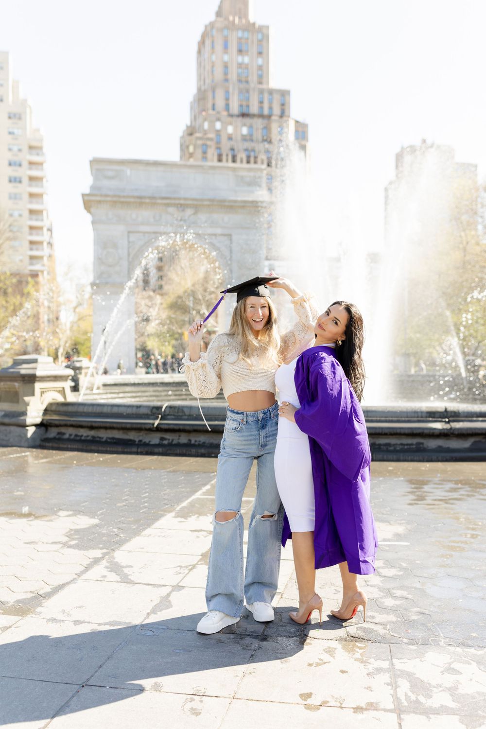 Two graduates celebrate at Washington Square Park fountain with the iconic arch and city buildings in background.