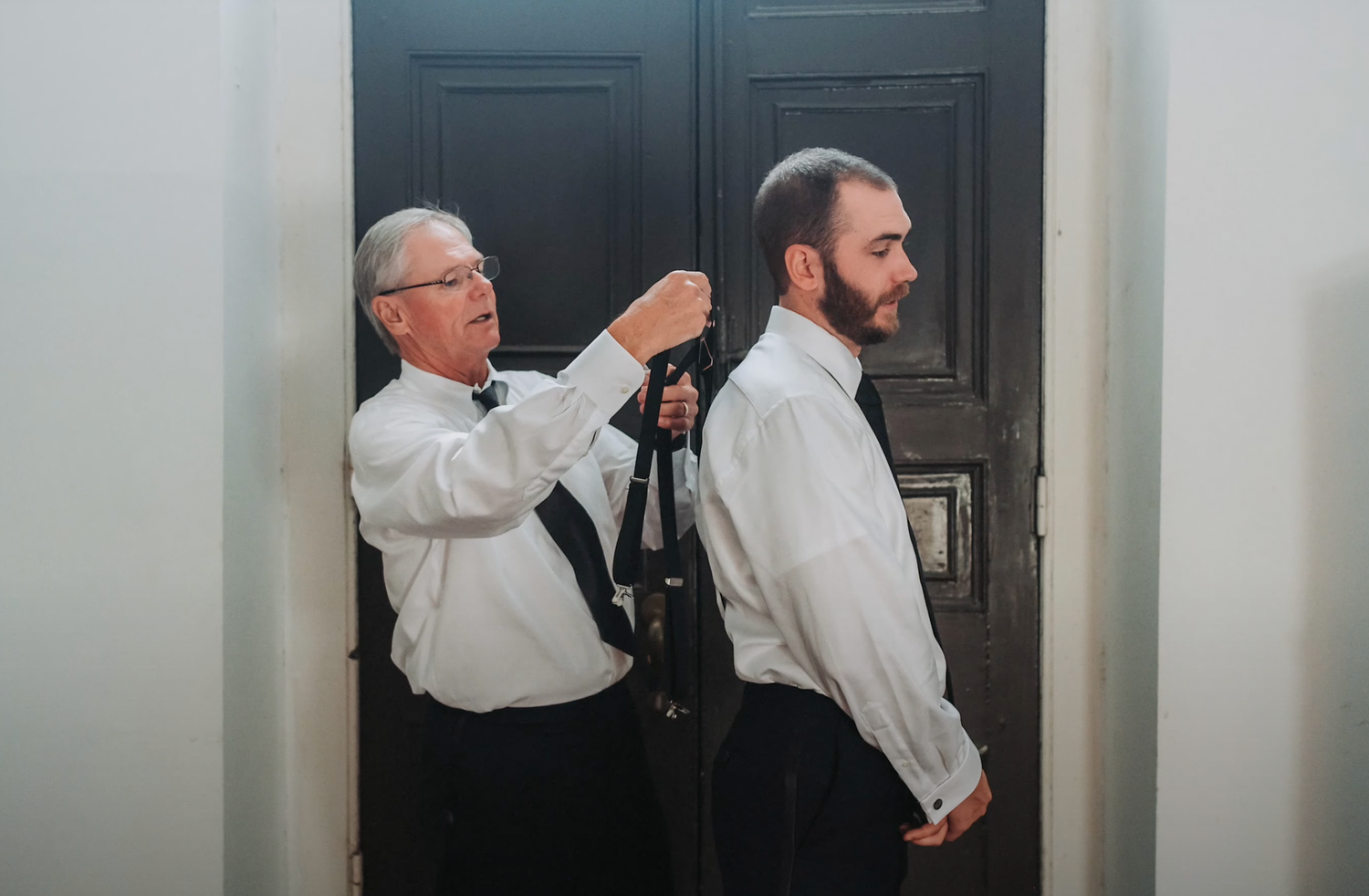 Two men in formal attire helping with wedding day preparations in a doorway.