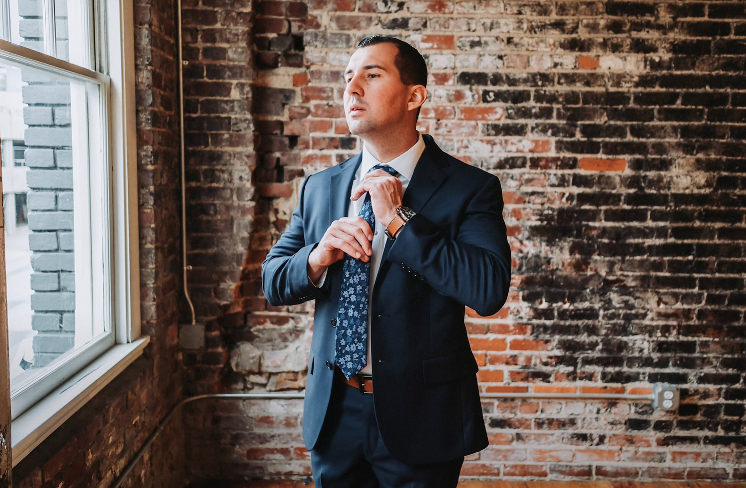 Man in navy suit adjusting his tie while standing next to brick wall and window.