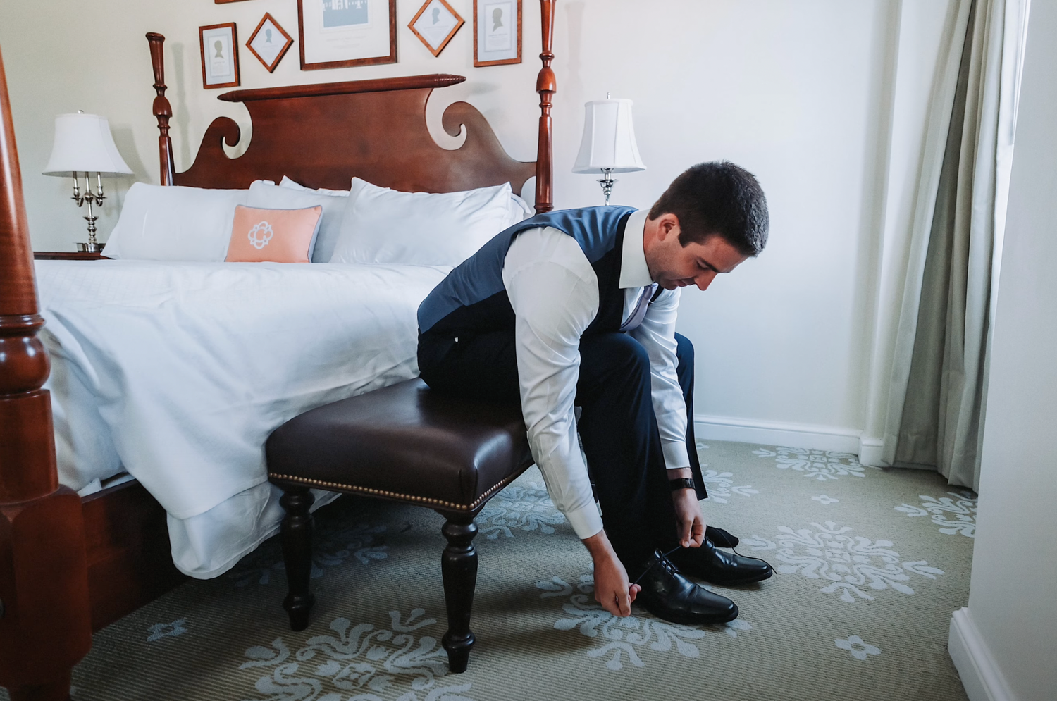Person in formal attire putting on dress shoes while sitting on bed in hotel room.