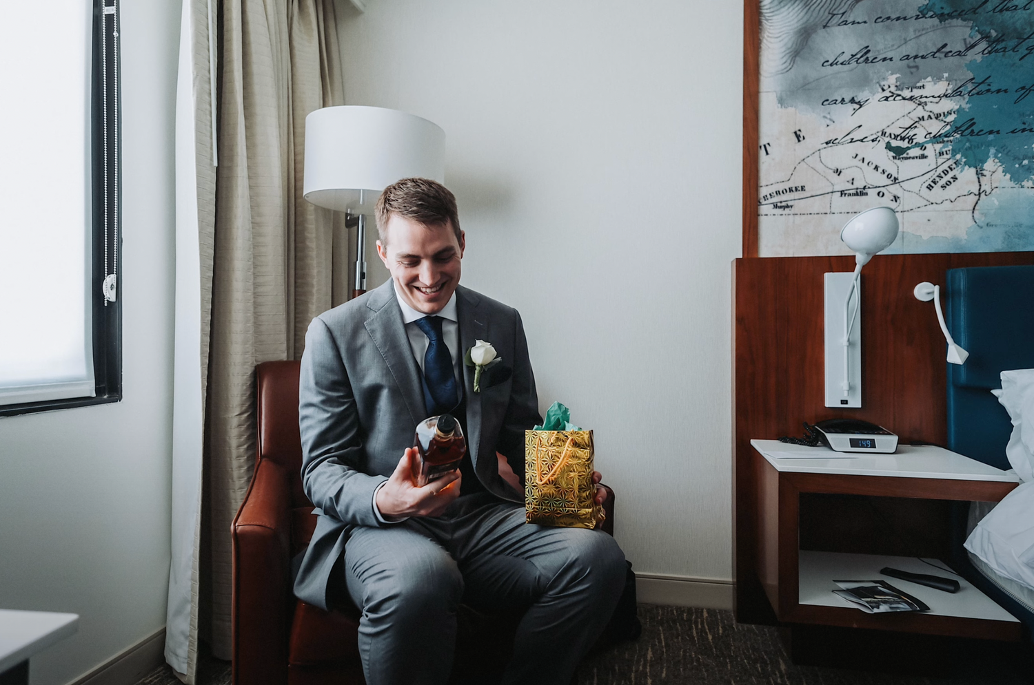 Person in gray suit sitting in hotel room chair next to window and lamp.
