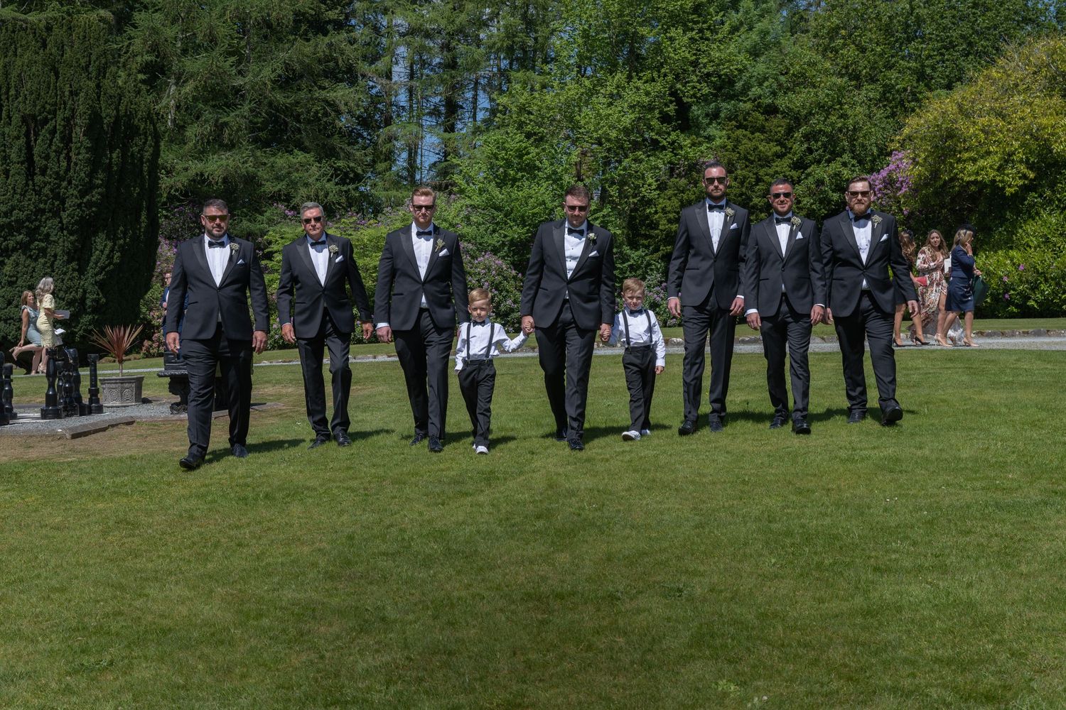 Groomsmen in black tuxedos walking together on grass during outdoor wedding.