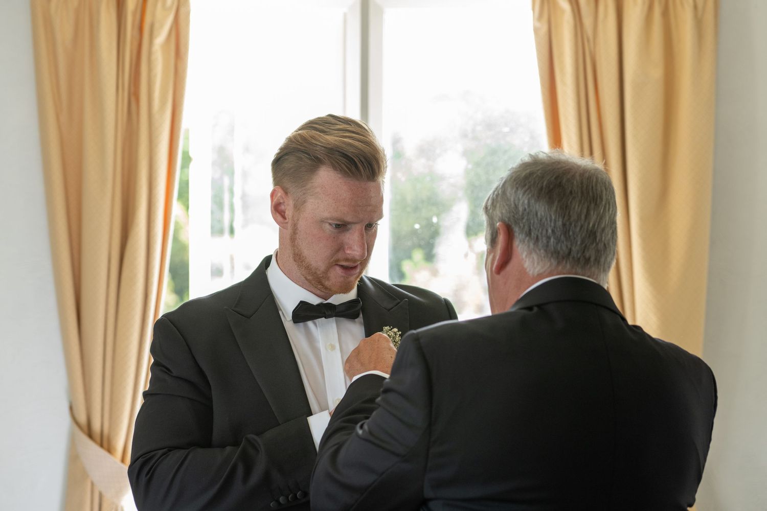 Two men in formal wear helping each other with bow ties by a window.