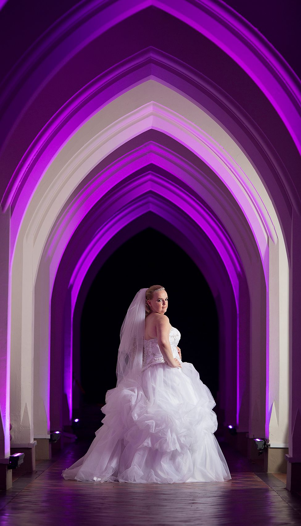 Bride in white wedding dress stands looking over her shoulder in the arched corridor of Gorton Monastery in Manchester
