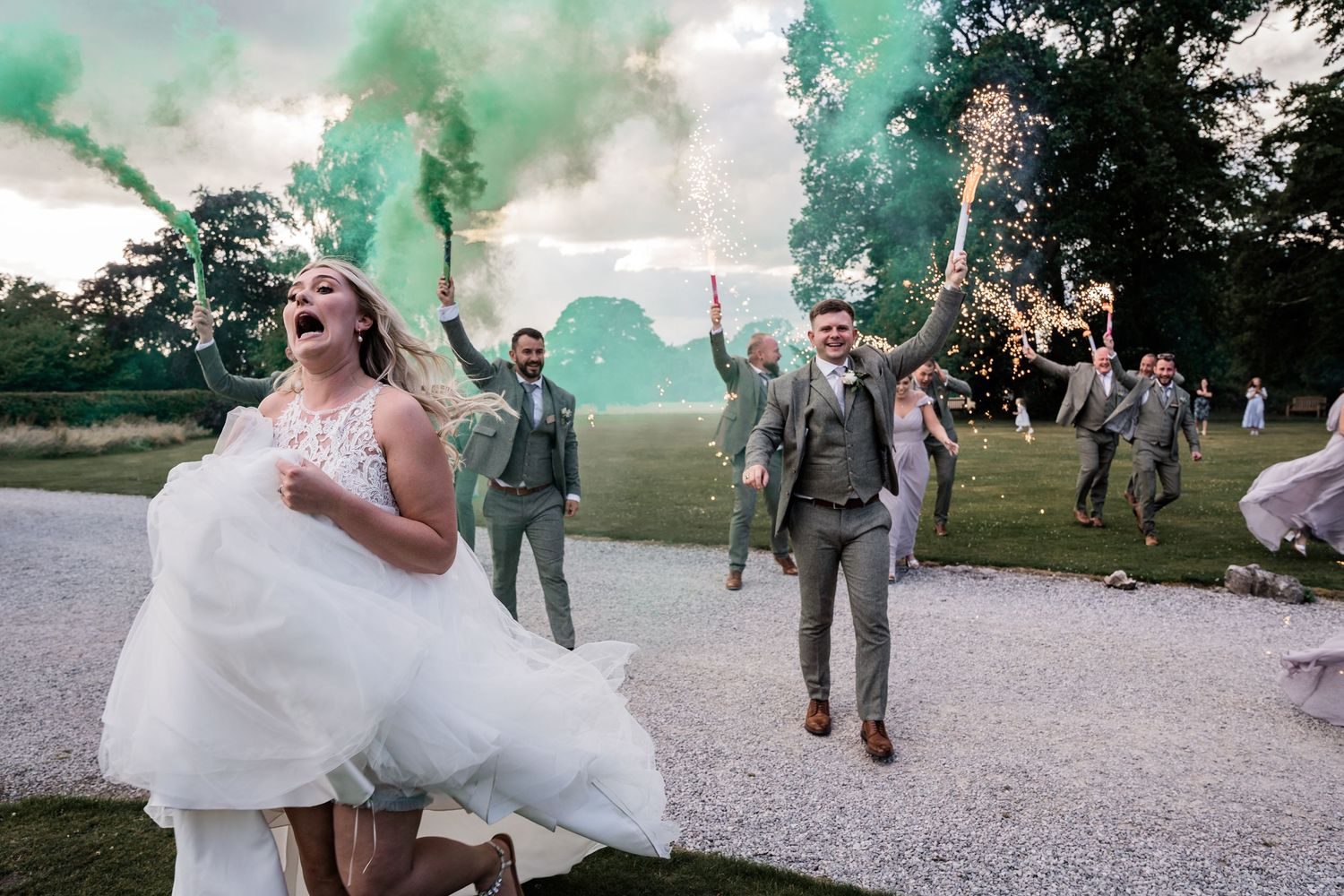 Wedding party celebration with colorful smoke bombs on gravel path.