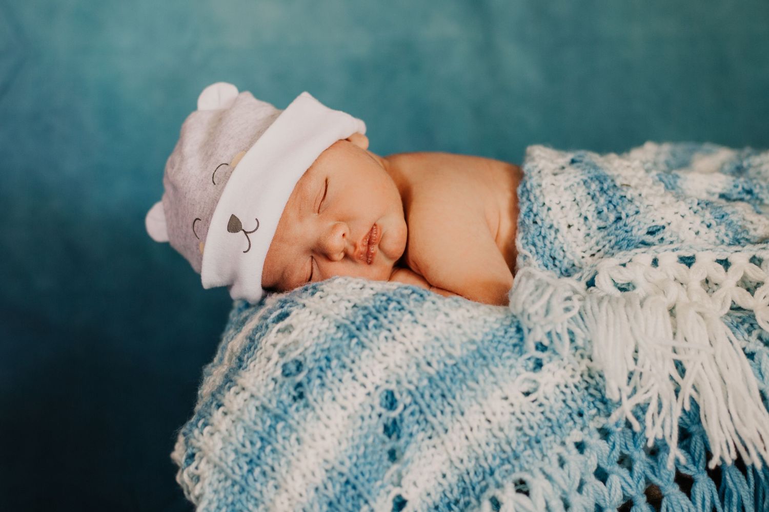 A sleeping newborn baby wearing a white cap on a blue and white crocheted blanket.