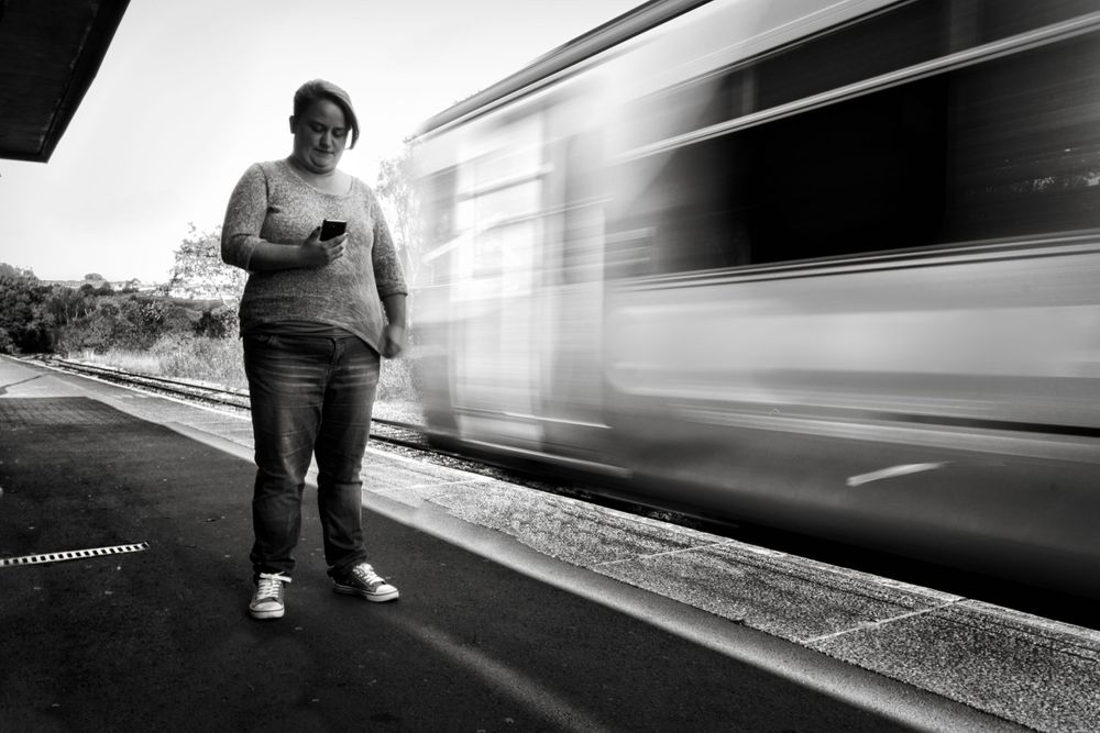 Person standing on train platform as blurred train passes in dramatic black and white composition.