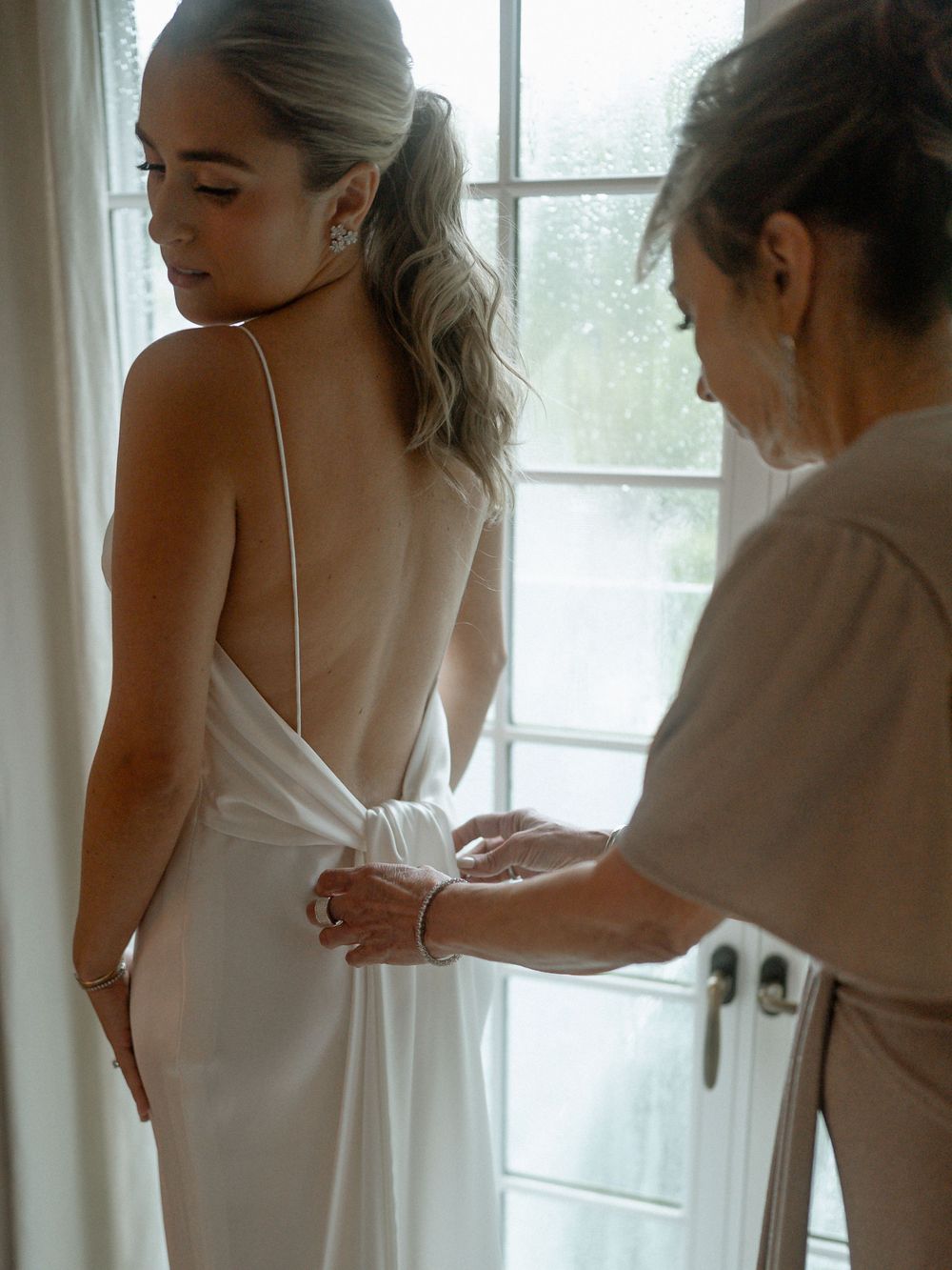 A stunning backless white wedding dress being fitted beside a bright window.