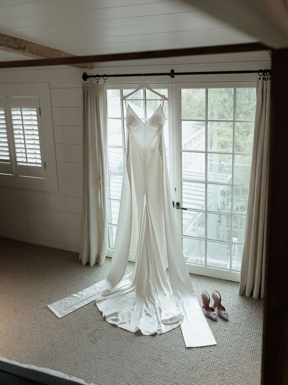 White wedding dress hangs by window in sunlit room with carpeted floor.