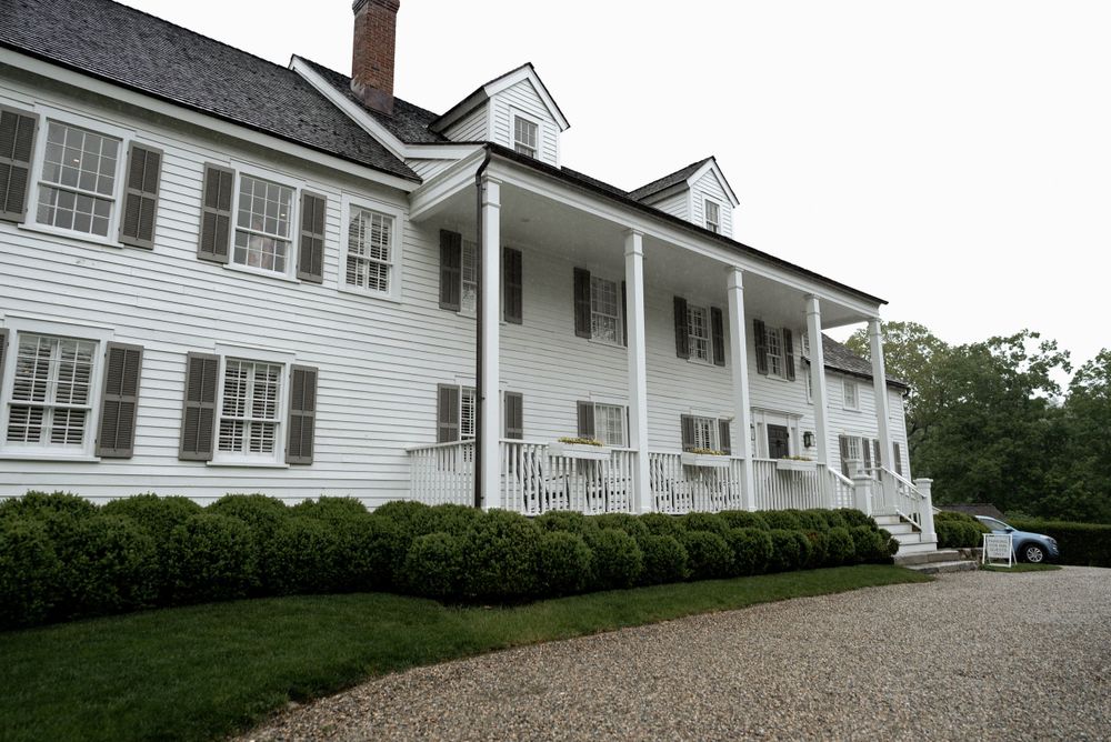 Classic white colonial mansion with columned porch and manicured hedges along gravel driveway.