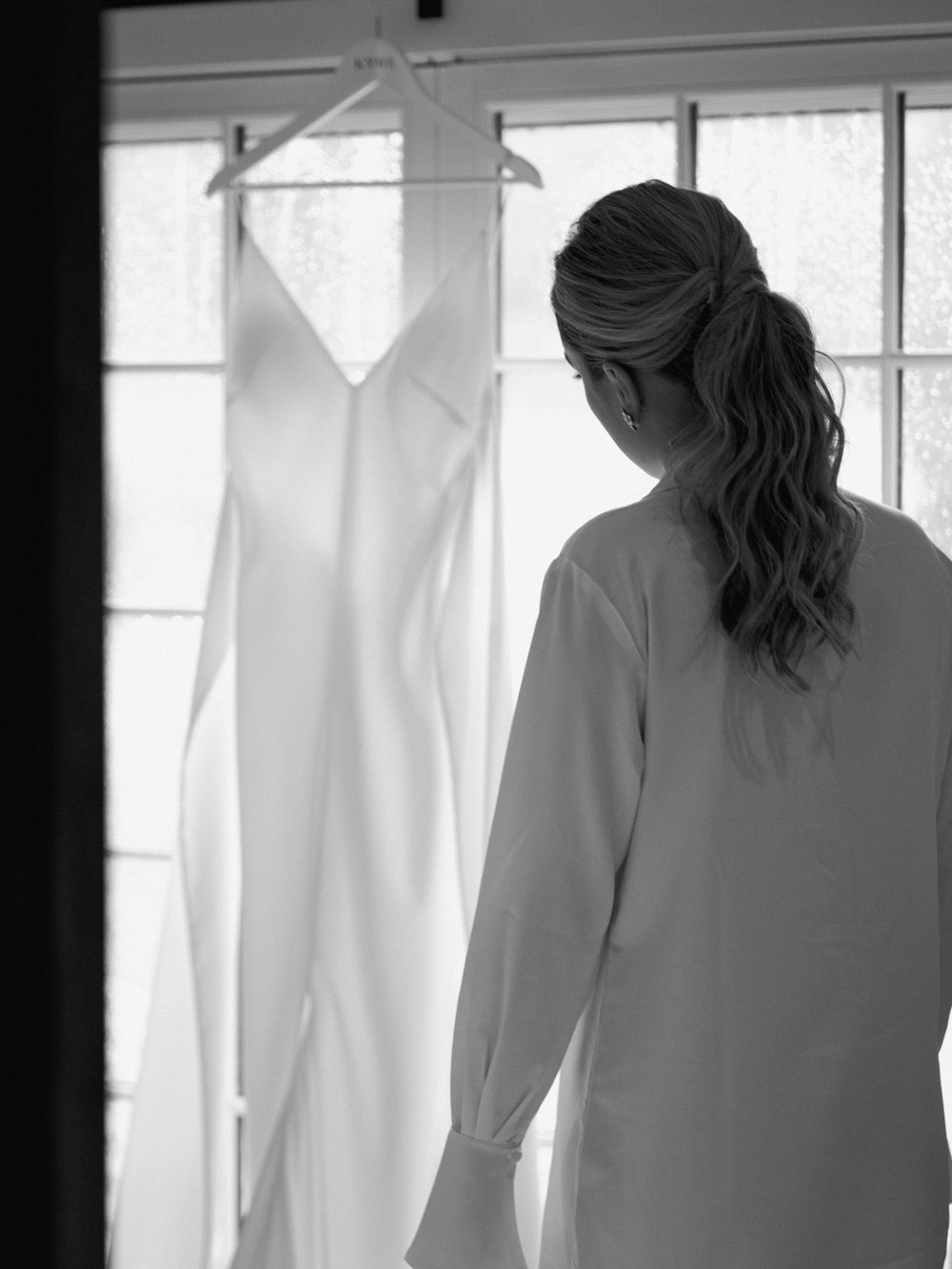 A sleek white wedding dress hangs by a window as a figure in white gazes at it in anticipation.