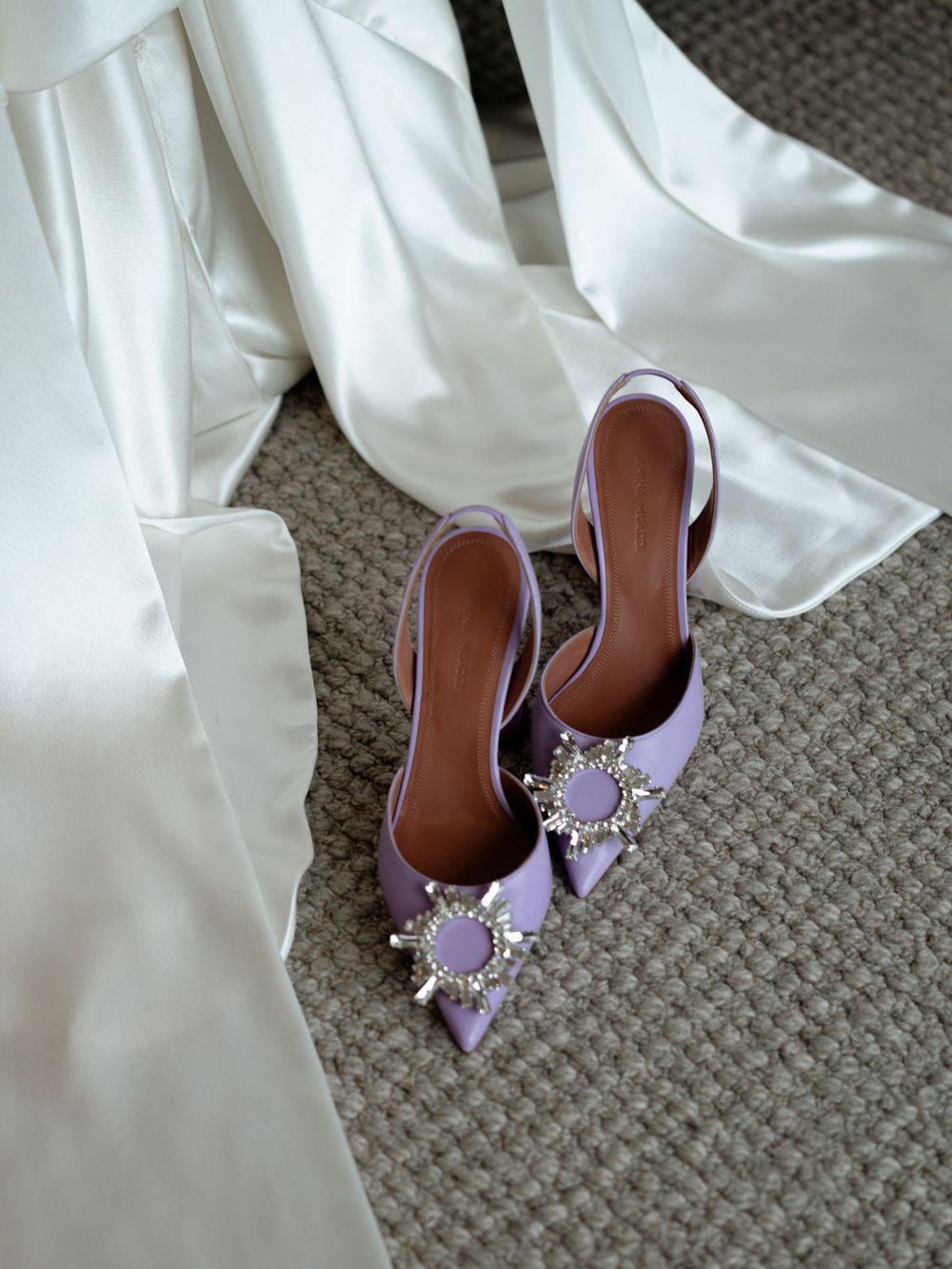 Purple embellished bridal shoes rest on carpet next to white fabric.