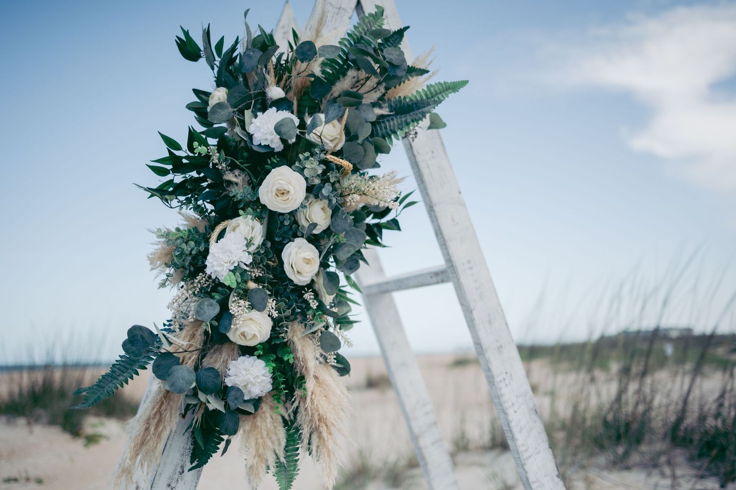 White and green floral arch arrangement against blue sky for beach wedding ceremony.