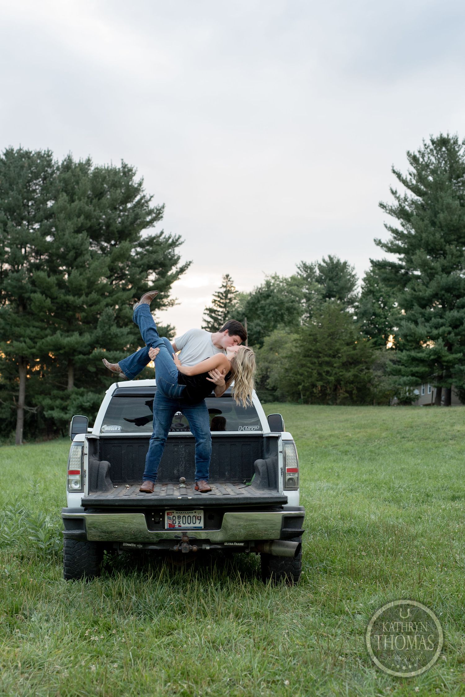A playful couple poses on top of their pickup truck in a field with tall pine trees.