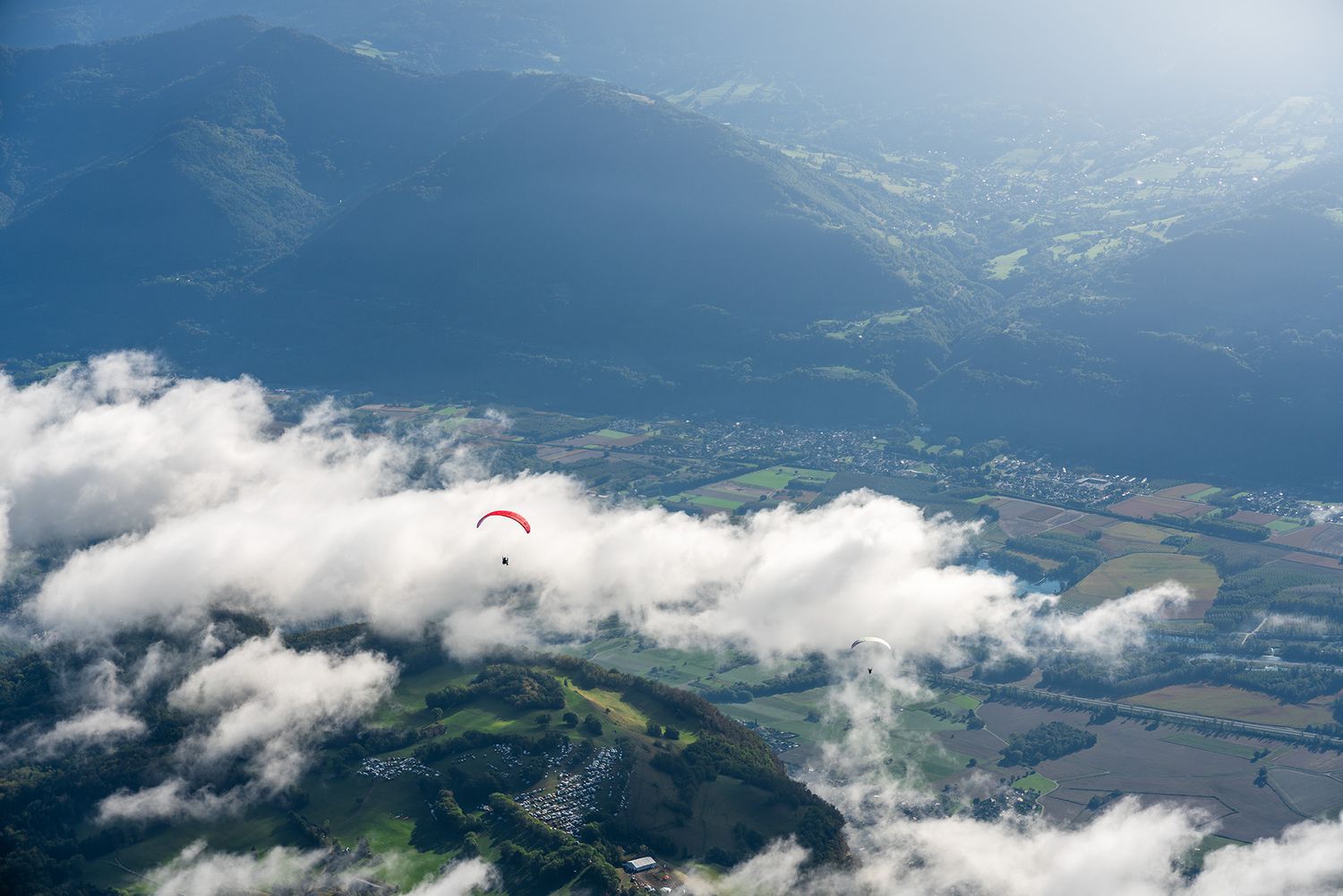 Paraglider soaring above clouds and mountains with lake valley visible below.