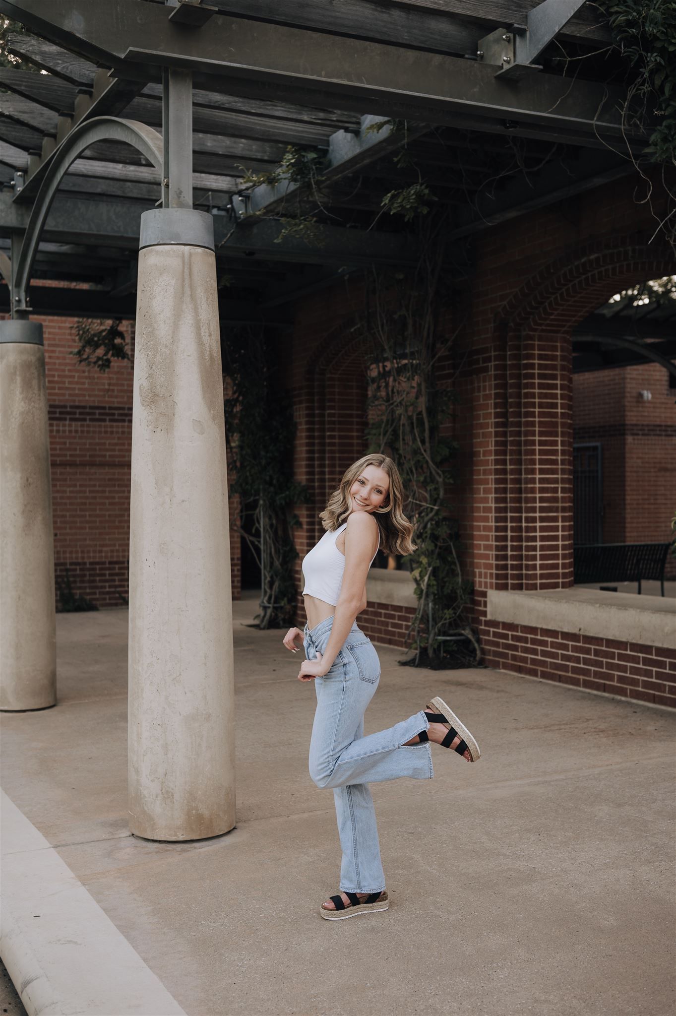Fashion model in casual outfit posing playfully against brick building with white columns.