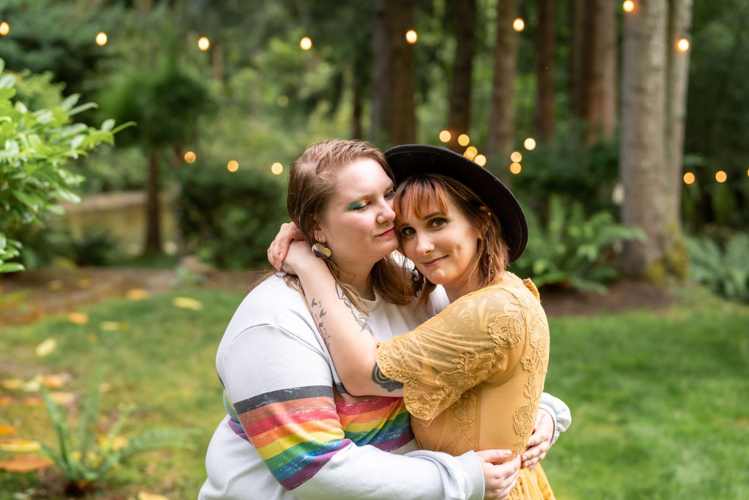 A couple embraces in a lush green garden setting with twinkling lights in the background.