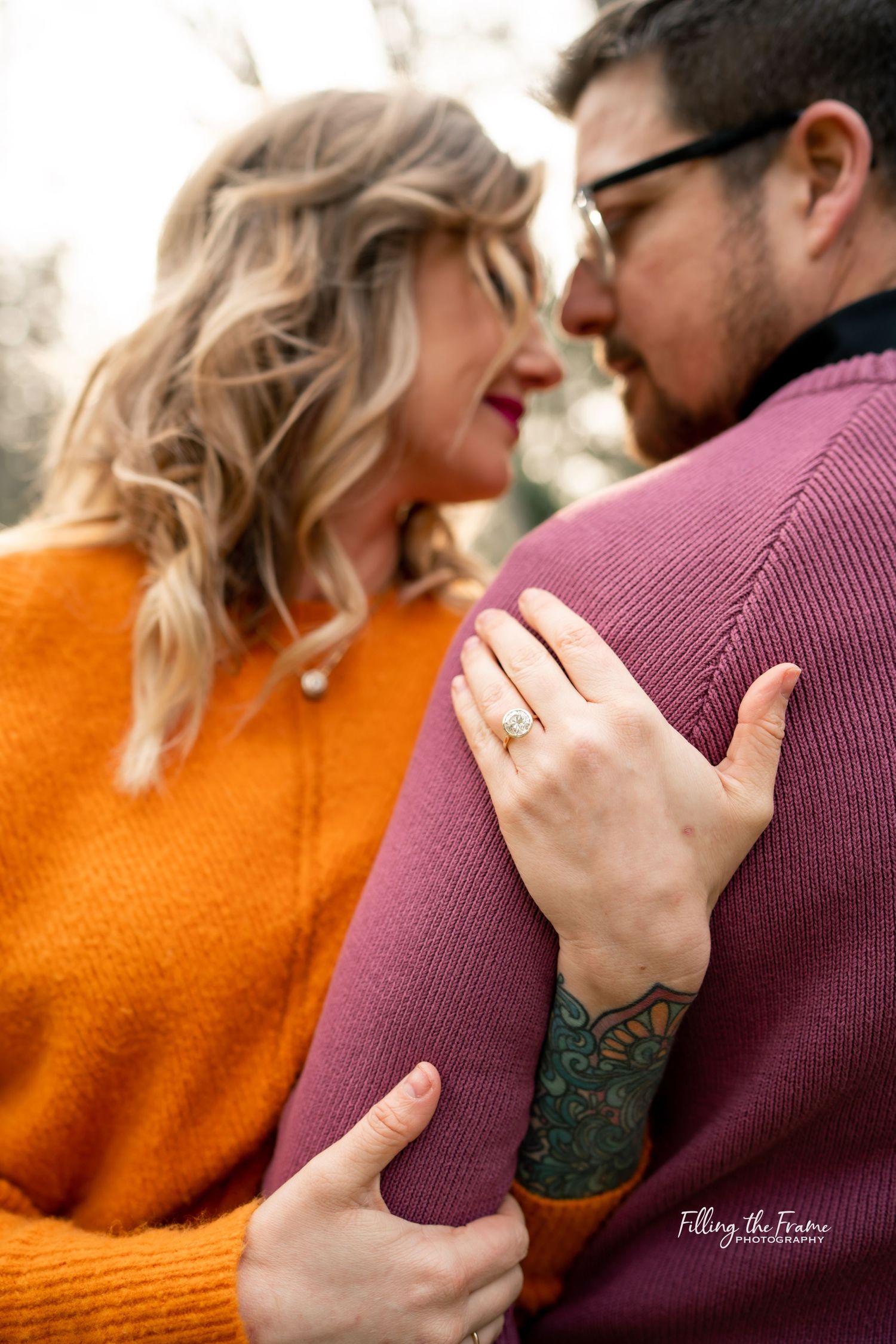 A close-up engagement photo showing an orange sweater, burgundy shirt, and a ring display.