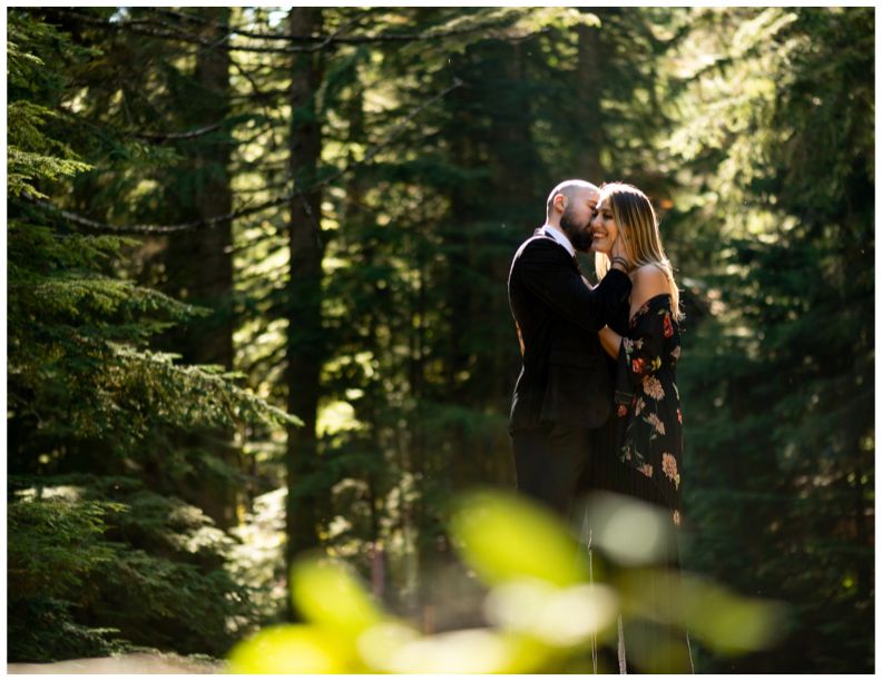 A romantic forest engagement photo with sunlight filtering through evergreen trees.