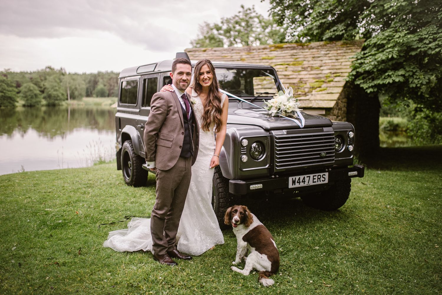 a couple stand by the lake at browsholme hall with a landrover and their spaniel dog