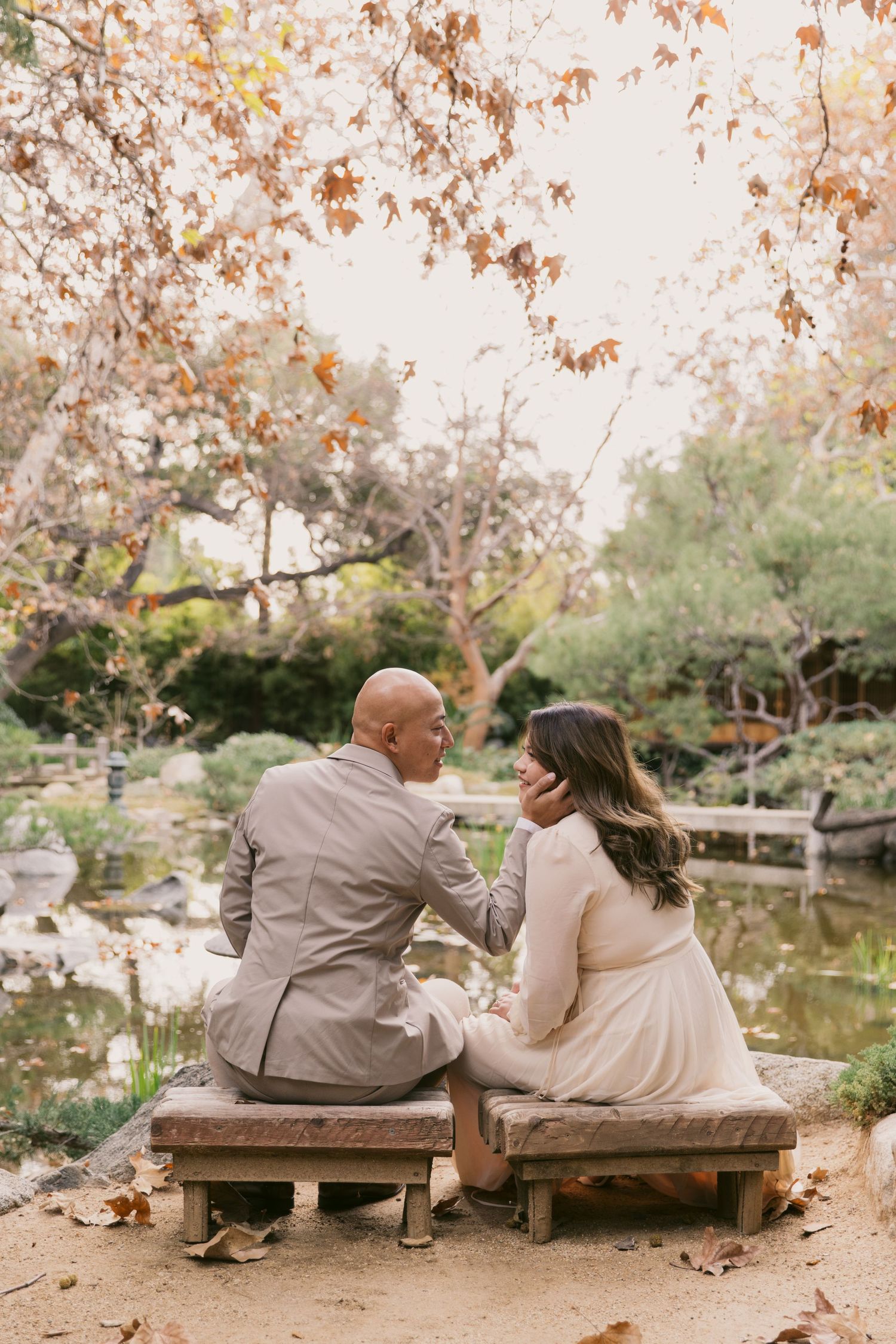 Couple sitting together on a bench near a pond surrounded by autumn foliage.