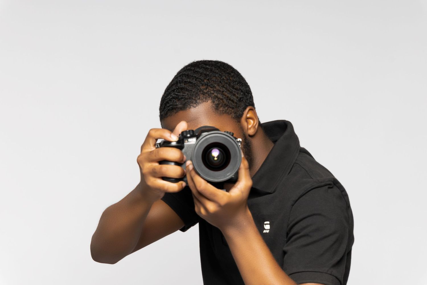 Photographer wearing black shirt holds DSLR camera against light gray background.